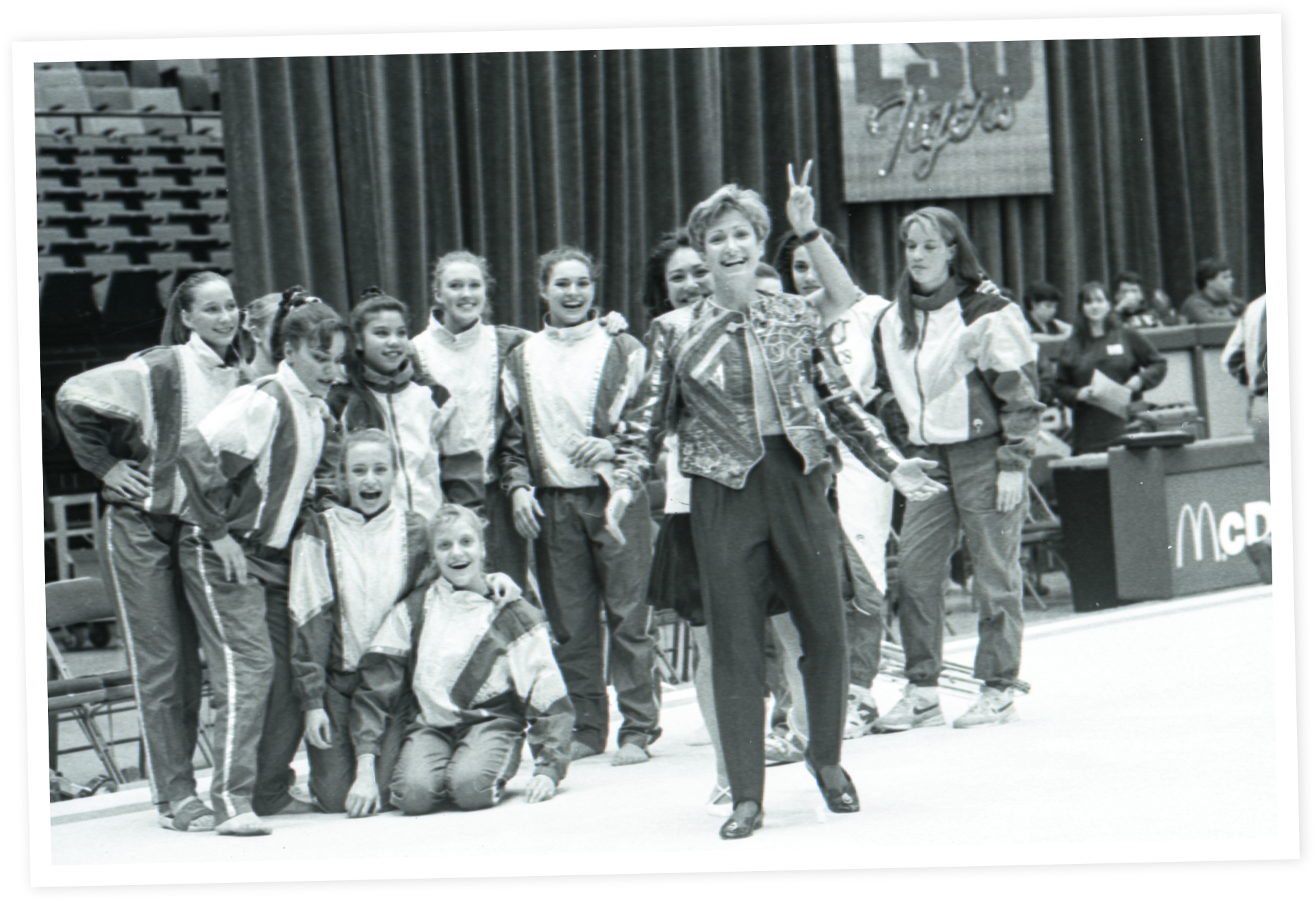 D-D in a patterned blazer and pants leading a group of young girls in track suits, in a gymnasium with a curtain in the background.