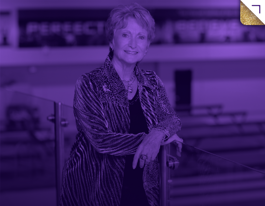 D-D smiling, wearing a patterned jacket and jewelry, standing with arms crossed in an indoor setting with a purple filter.