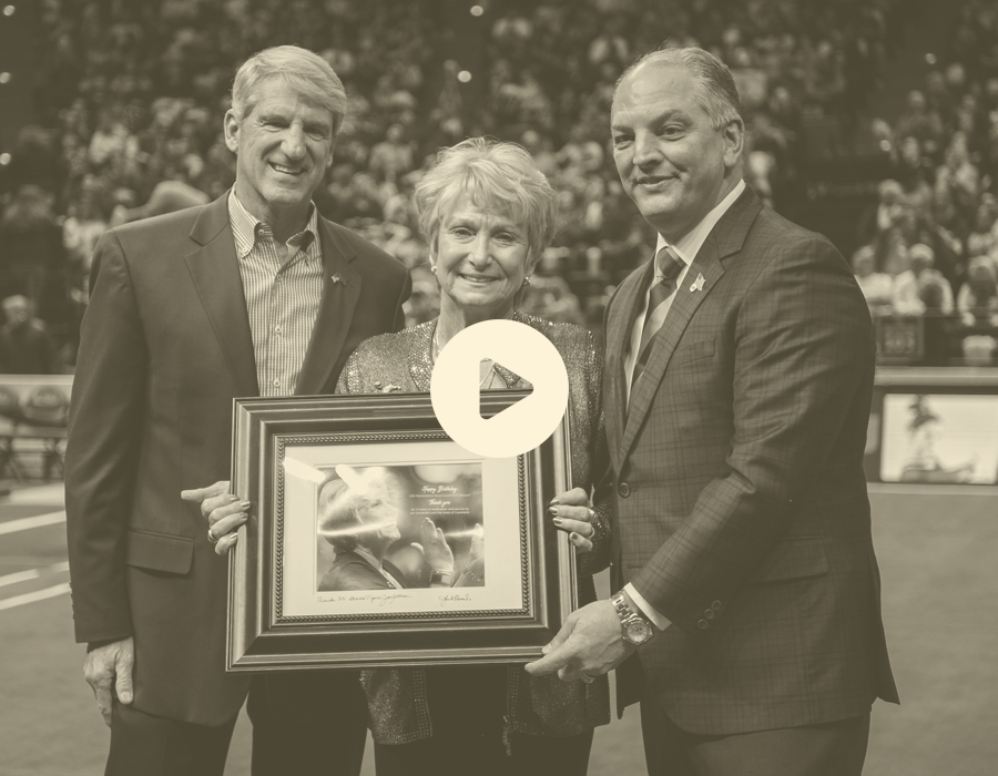Three people, two men and one woman, standing on a sports field at a stadium, holding a framed picture and smiling at the camera.