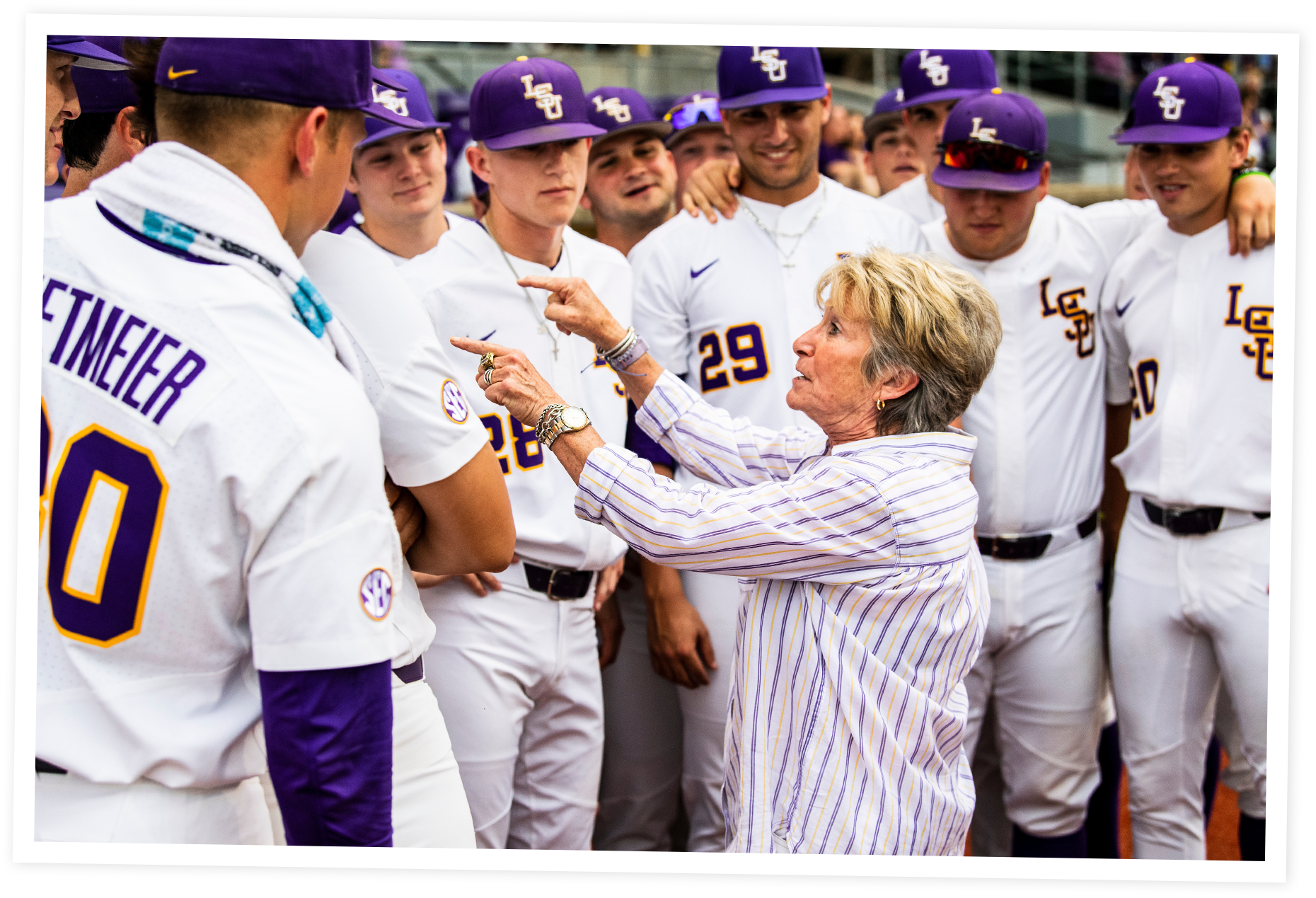 D-D Breaux in a striped shirt is talking and gesturing towards a group of baseball players in purple and white uniforms during a game or practice. The players are standing close together, and some are wearing purple hats and sunglasses.