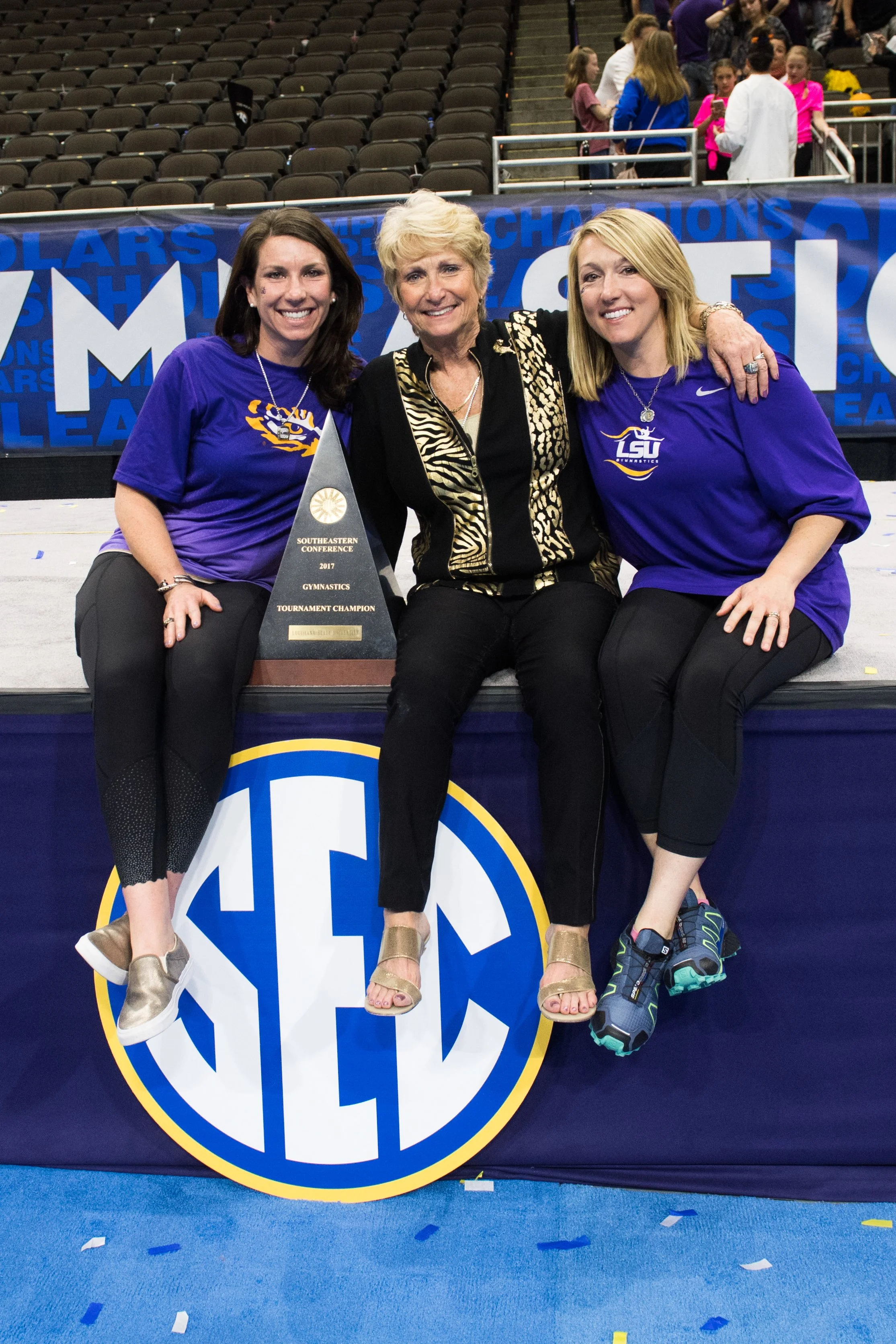 Three women sitting on a table with an SEC sign, celebrating a gymnastics tournament championship with a trophy, in an indoor arena with spectators in the background.