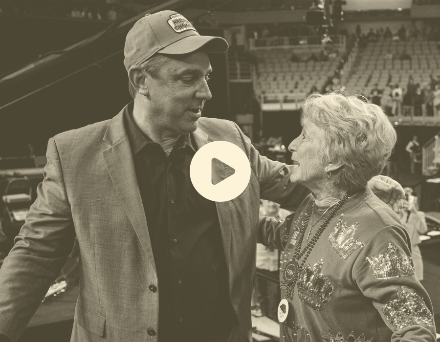 A man and woman are talking in a large indoor sports arena, standing close to each other and smiling. The man is wearing a baseball cap and blazer, and the woman is wearing a decorated sweater with a necklace.