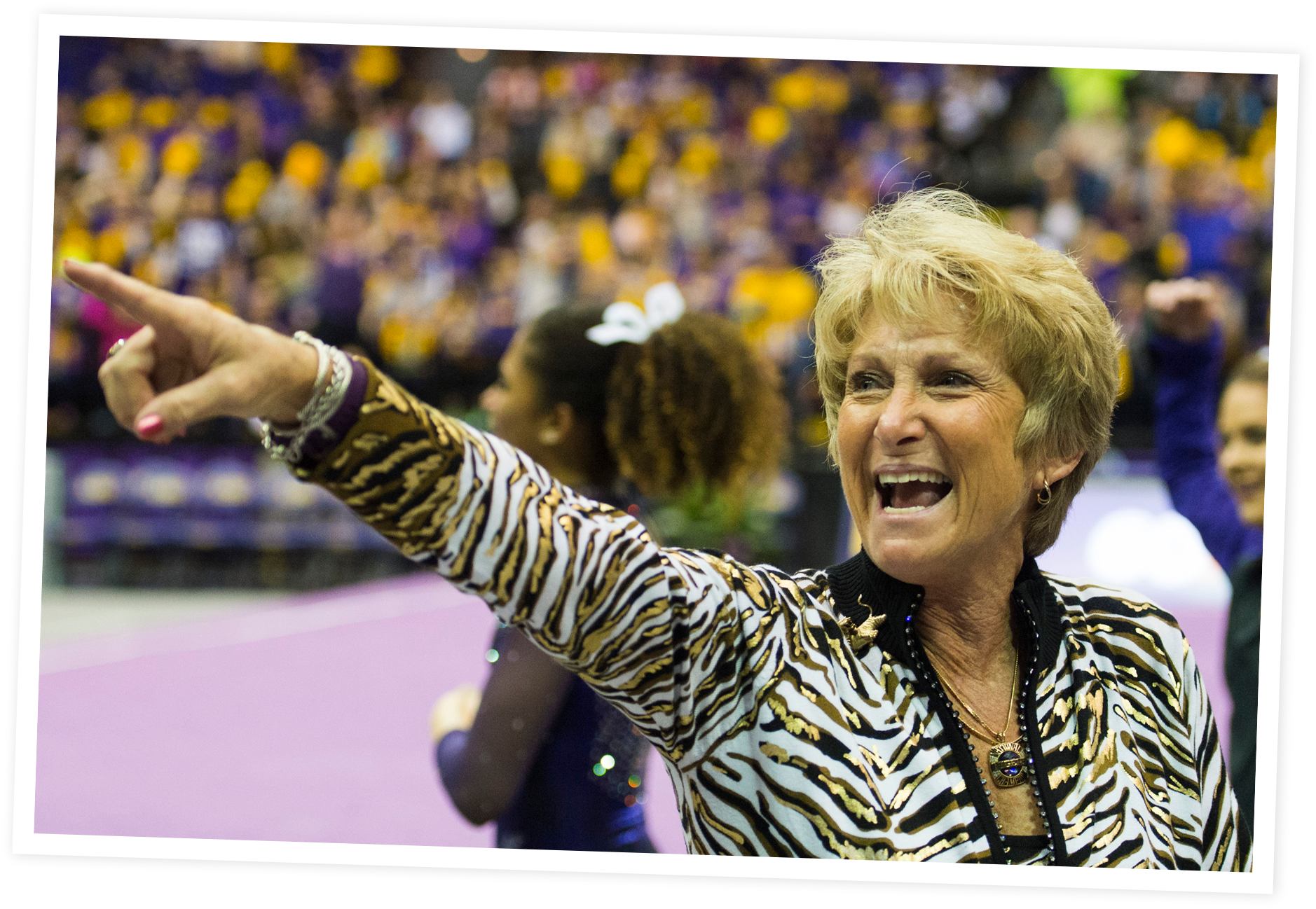 D-D Breaux with short blonde hair, wearing a zebra print jacket and jewelry, is pointing and shouting towards a sports event with a crowd of spectators in the background.