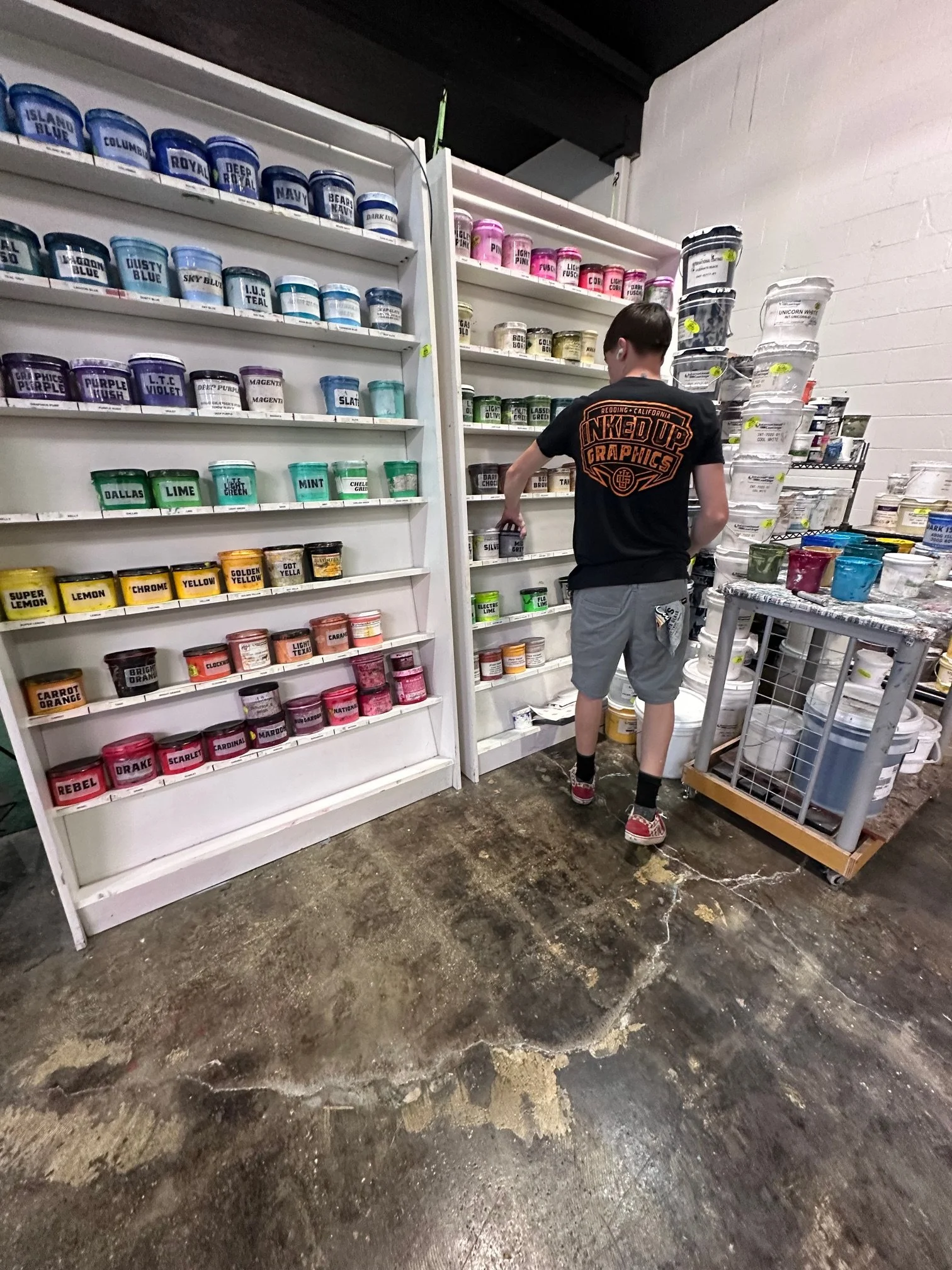 A person with short hair wearing a black t-shirt and gray shorts is shopping for paint in a store, facing shelves filled with various colors of paint containers.