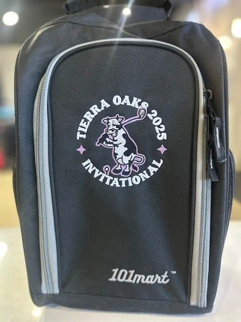 A black backpack with an embroidered circular logo featuring a tiger holding a lollipop, with the words "Tierra Oaks 2025 Invitational" around it. The logo is purple and white, and the backpack has a gray zipper and a logo of 10lmart in the lower corner.
