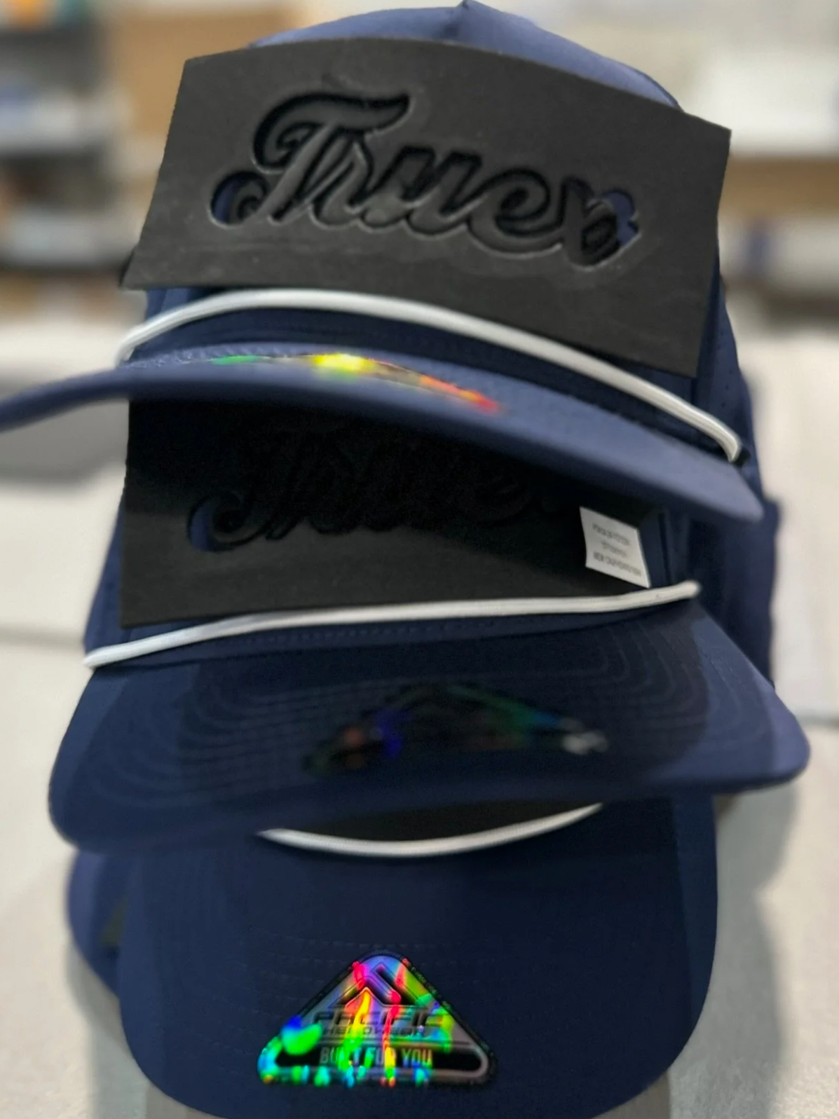 A stack of blue caps, with a large black tag that reads 'Trucker' on top, and a holographic sticker featuring a rainbow-colored triangle and the text 'Built for You' at the bottom.