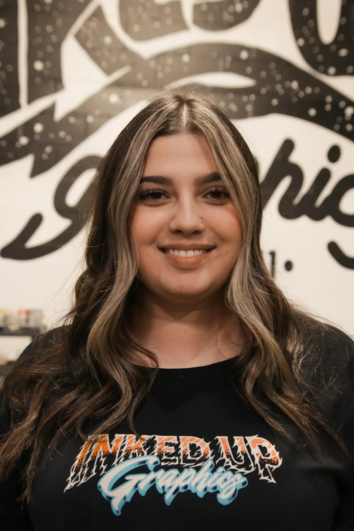 A young woman with long, wavy hair smiling at the camera, standing in front of a black wall with white artistic lettering, wearing a black T-shirt with colorful graphic text reading 'INKED up Graphics'.