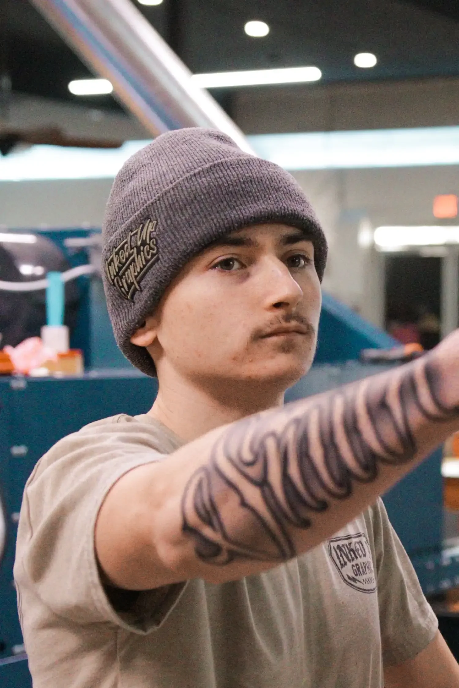 A young man wearing a gray knit beanie and beige t-shirt with a tattoo on his arm, reaching out with his arm extended. The background appears to be an indoor public space, possibly a store or restaurant.
