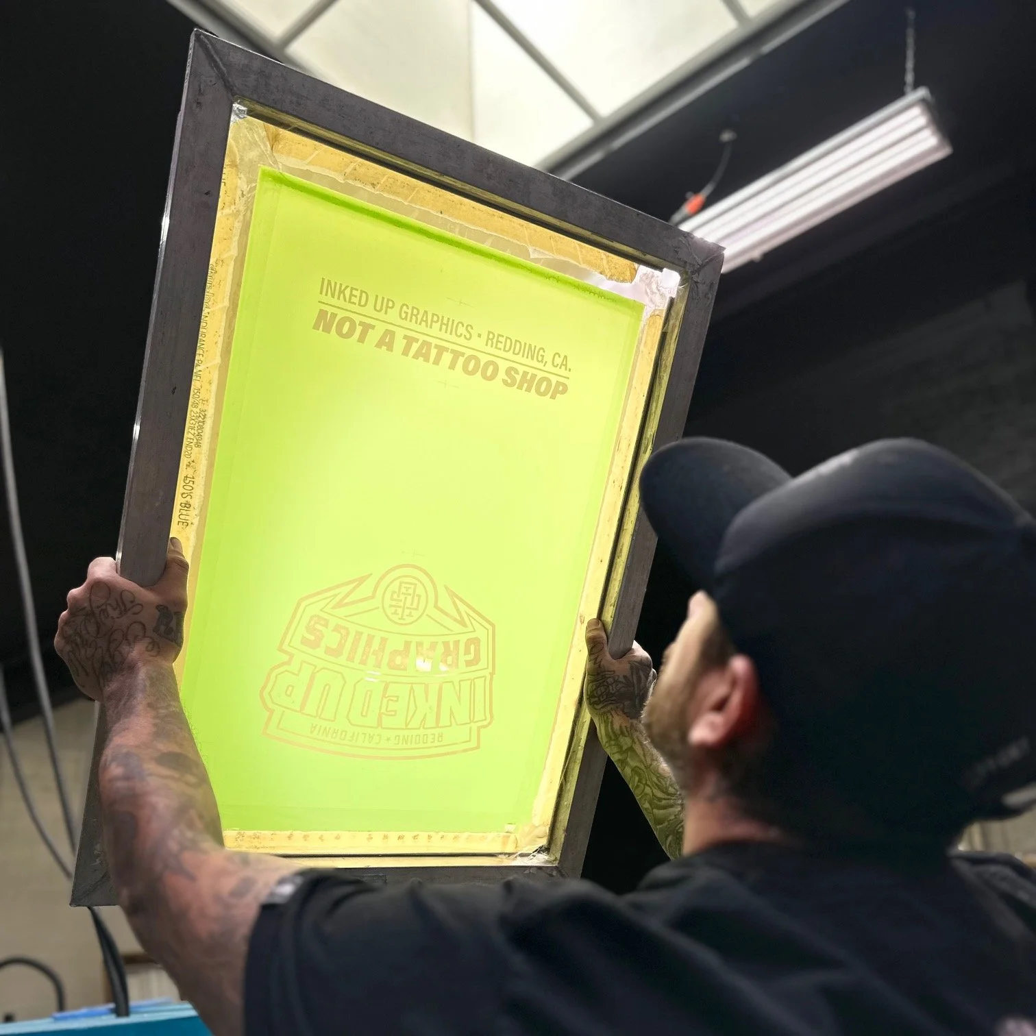 Person holding a yellow and green sign with text reading, 'INKED UP GRAPHICS - REDDING, CA. NOT A TATTOO SHOP', with tattooed arms and a tattooed head.