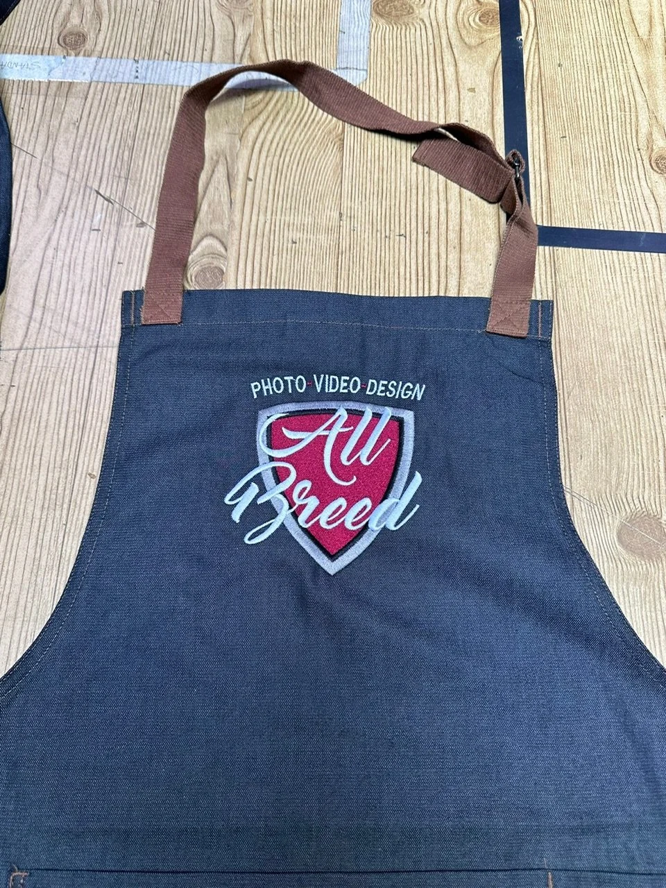 A dark apron with a patch that says 'All Breed' and 'Photo Video Design' on a wooden surface.