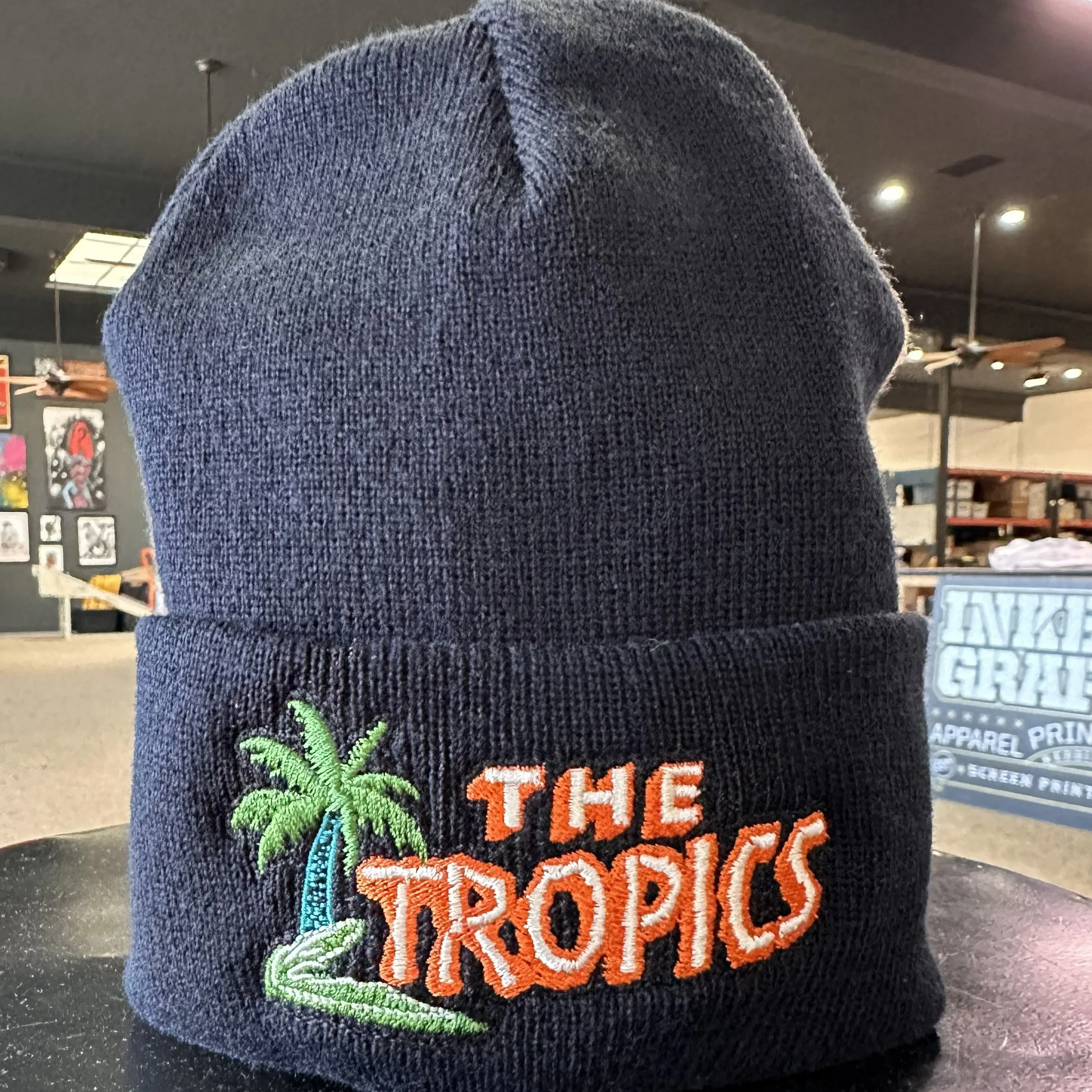 A beanie hat with embroidered design of a green cannabis leaf and the text 'The Reaper' in orange and white letters. The background is a dark indoor setting with some posters on the wall.
