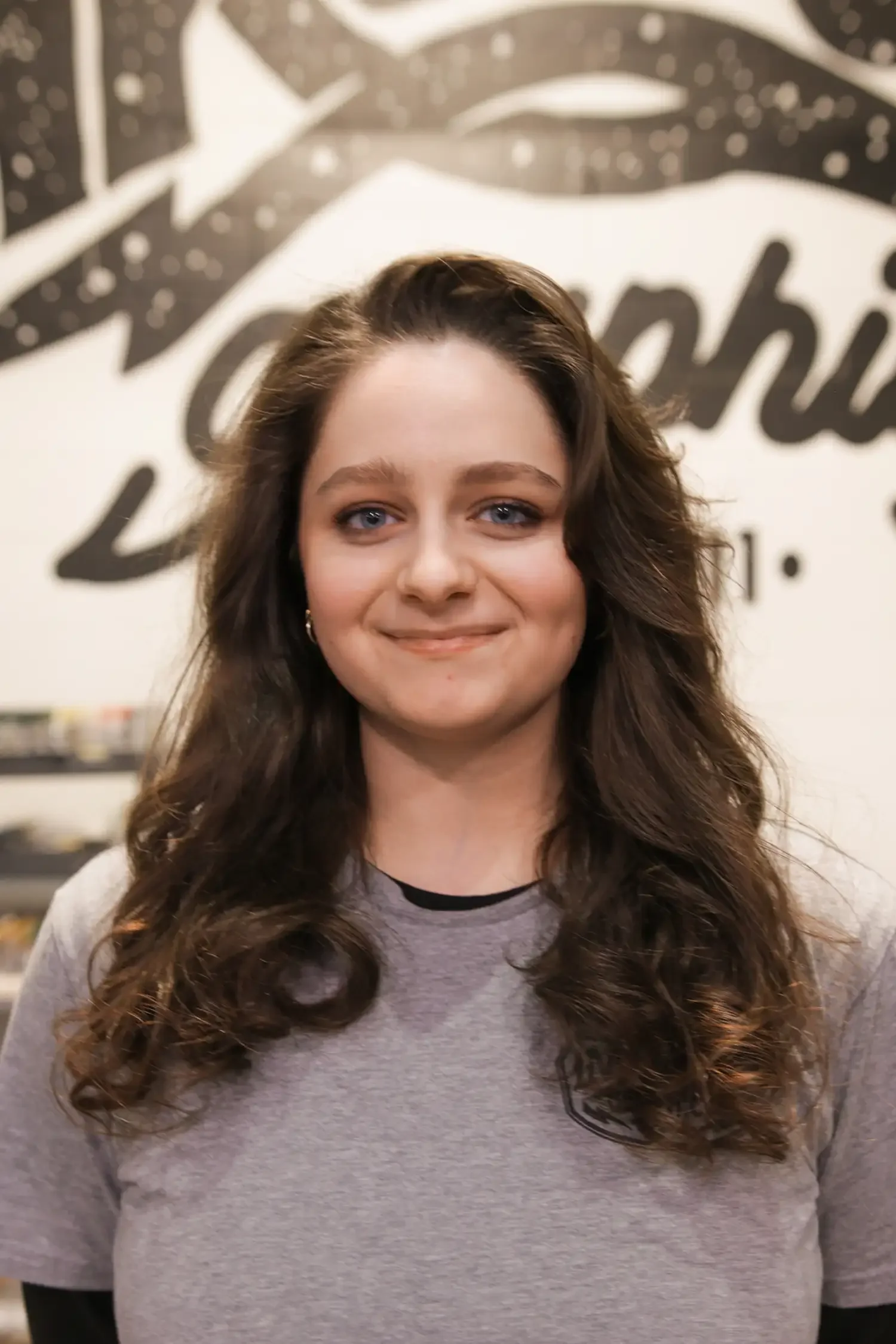 A young woman with long, wavy brown hair and blue eyes, smiling, wearing a gray t-shirt, in front of a wall with black and white artistic writing.