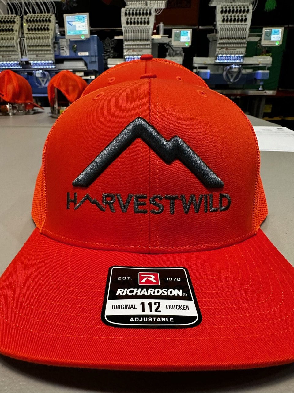 A red baseball cap with black embroidered mountain graphic and the words 'HARVESTWILD' on the front, displayed on a store counter with embroidery machines in the background.