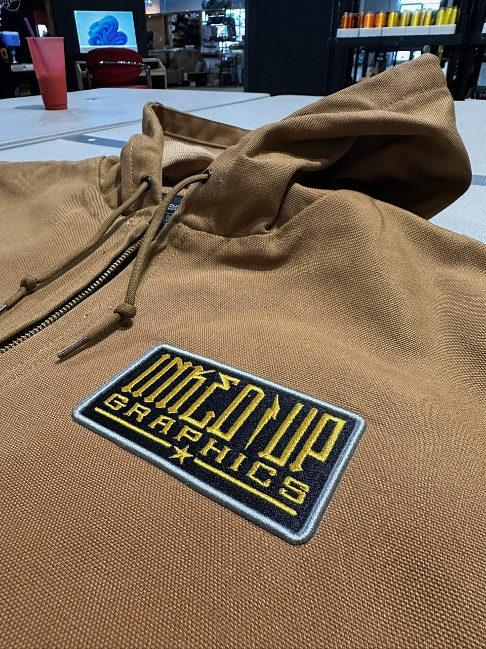Close-up of a tan hoodie with a 'Clinton Brothers Graphics' patch on the chest, on a table in a store or cafe setting.