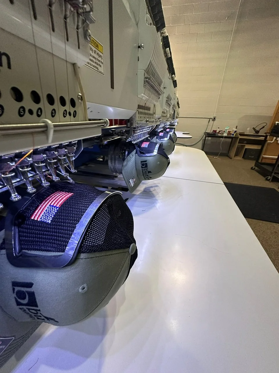 Close-up of a multi-head embroidery machine stitching an American flag design onto a cap, with a workspace in the background.