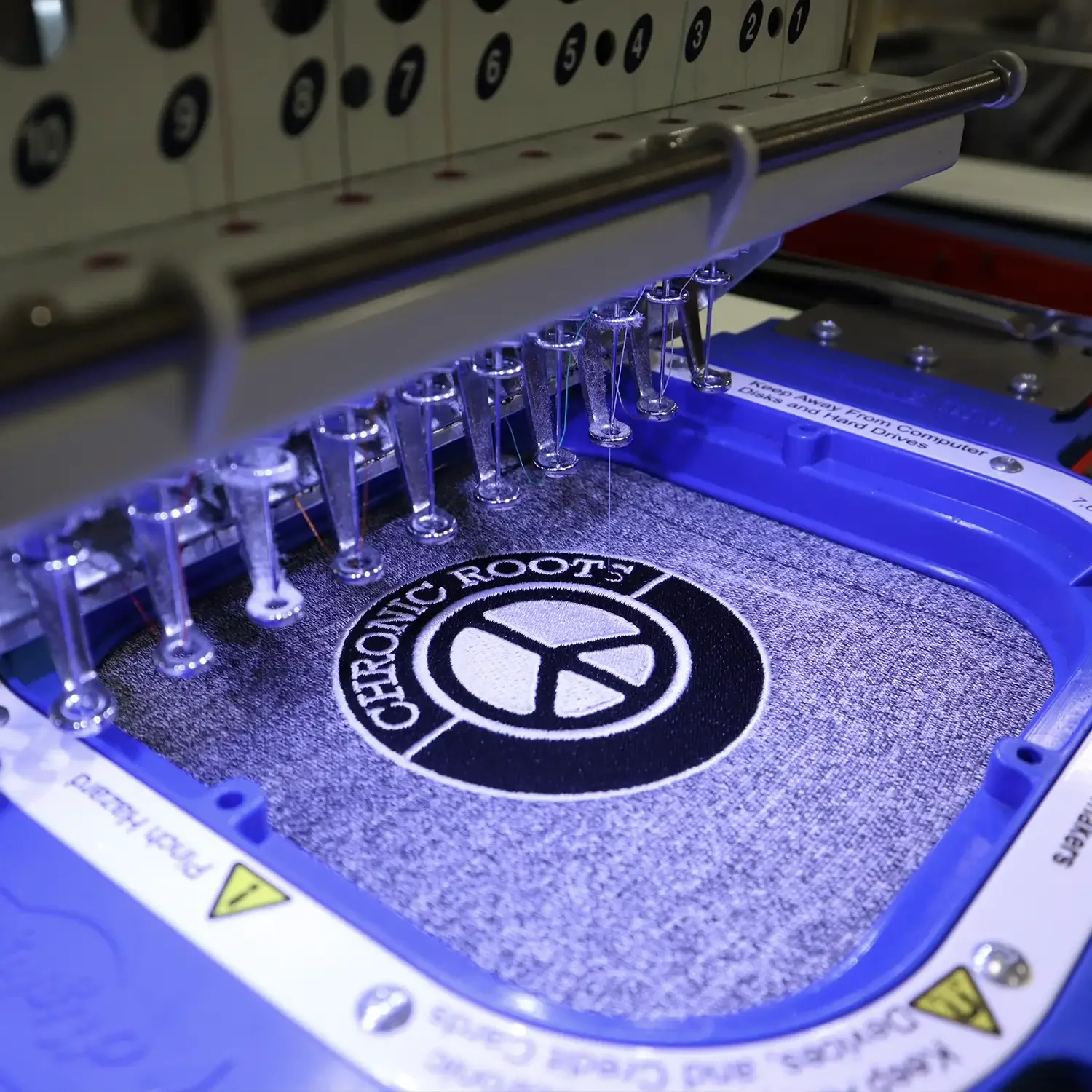 Embroidery machine stitching a black and white circular patch with the Peace symbol and the words "CHRONIC ROOTS" around it.