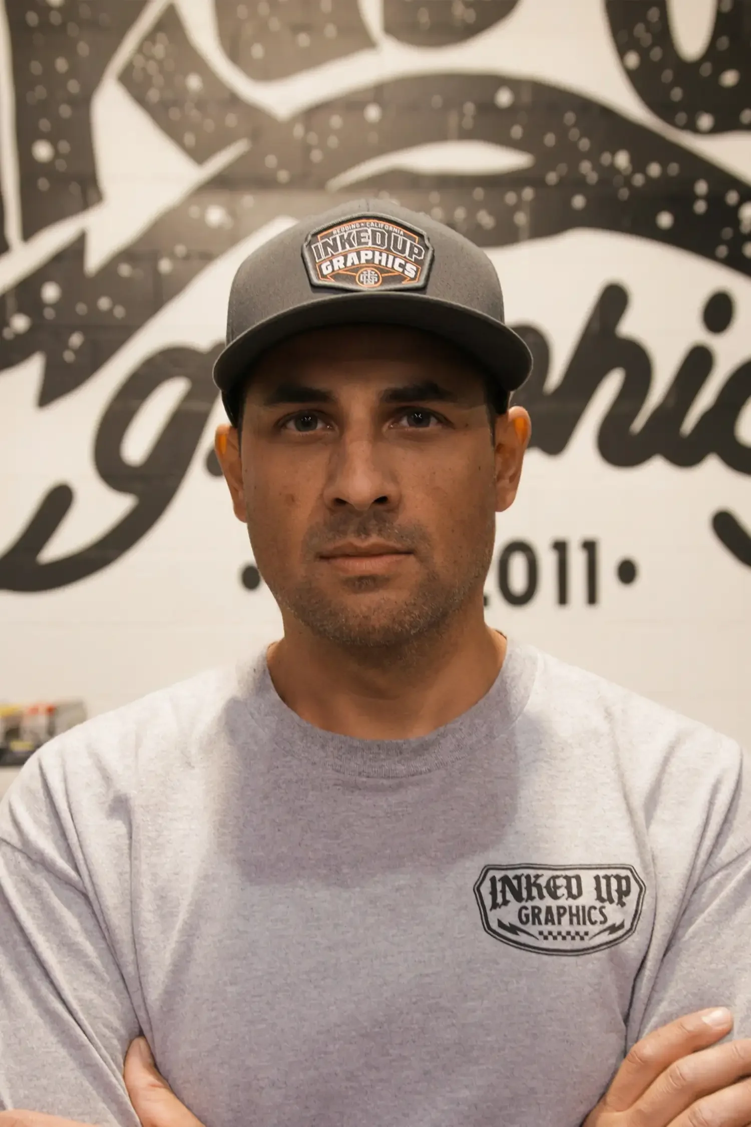 A man wearing a gray baseball cap and a gray t-shirt with 'Inked Up Graphics' logo poses in front of a mural with the same logo.