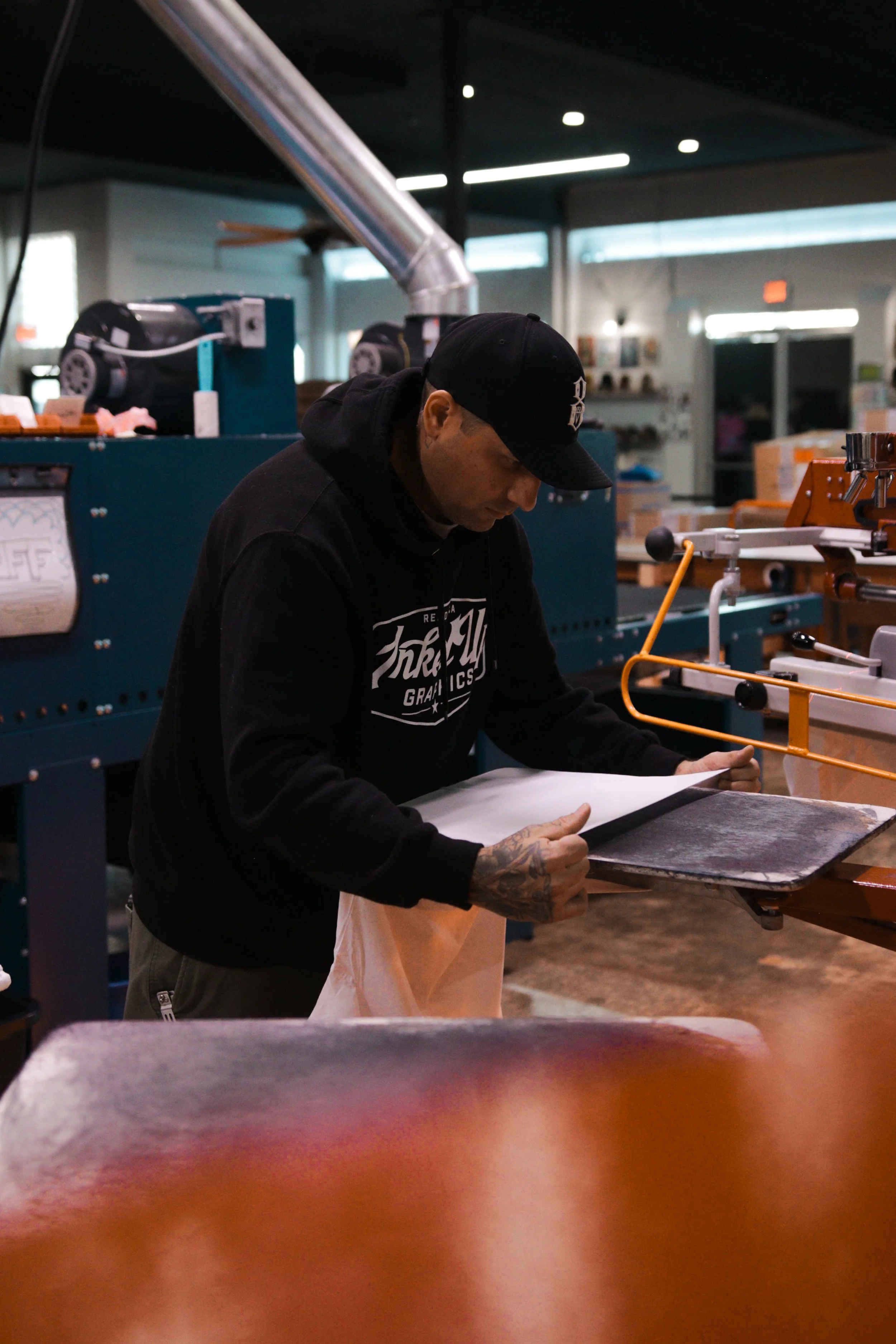 A man with tattoos on his right hand wearing a black hoodie and a black cap, working in a workshop or factory setting. He is looking down at some material on a table, with industrial machinery and various tools in the background.