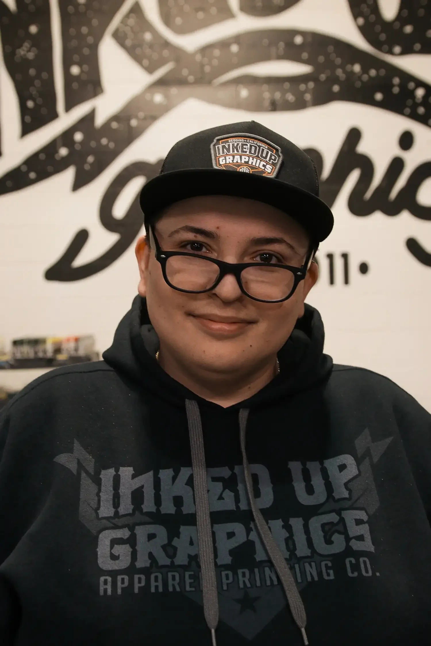 A person smiling at the camera, wearing glasses, a black hoodie with the text "LIKED UP GRAPHICS APPAREL PRINTING CO." and a black cap with the logo "Inked Up Graphics" in front of a wall with a graphic design.