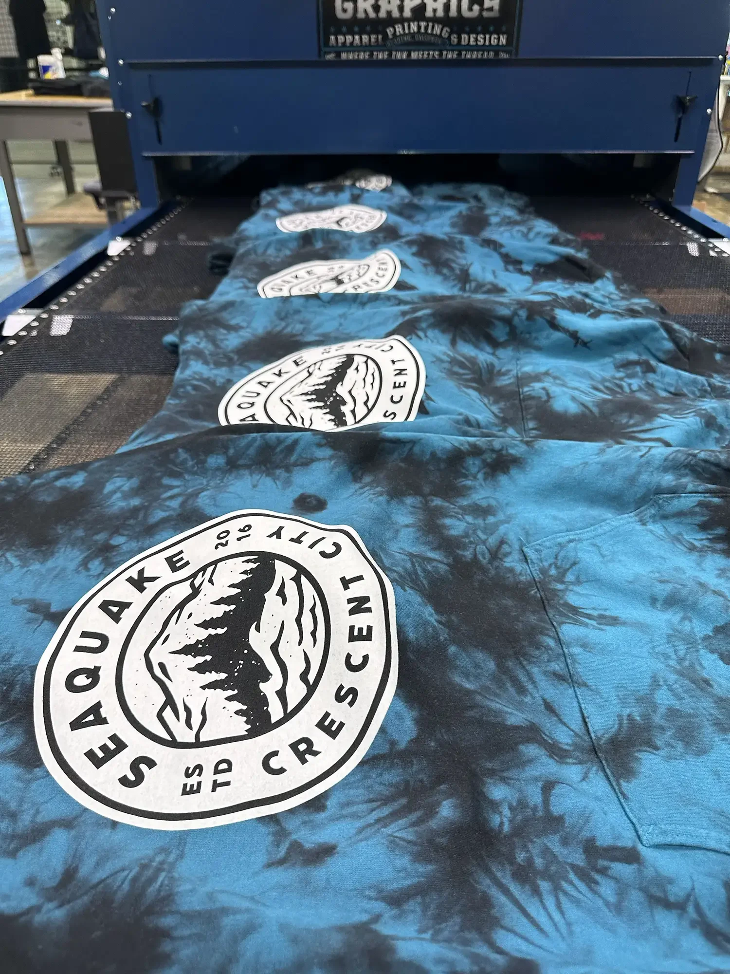 A large blue tie-dye banner with multiple black and white logos for 'Eauke City 2018 EST. SCENT' on a printing machine in a workshop.