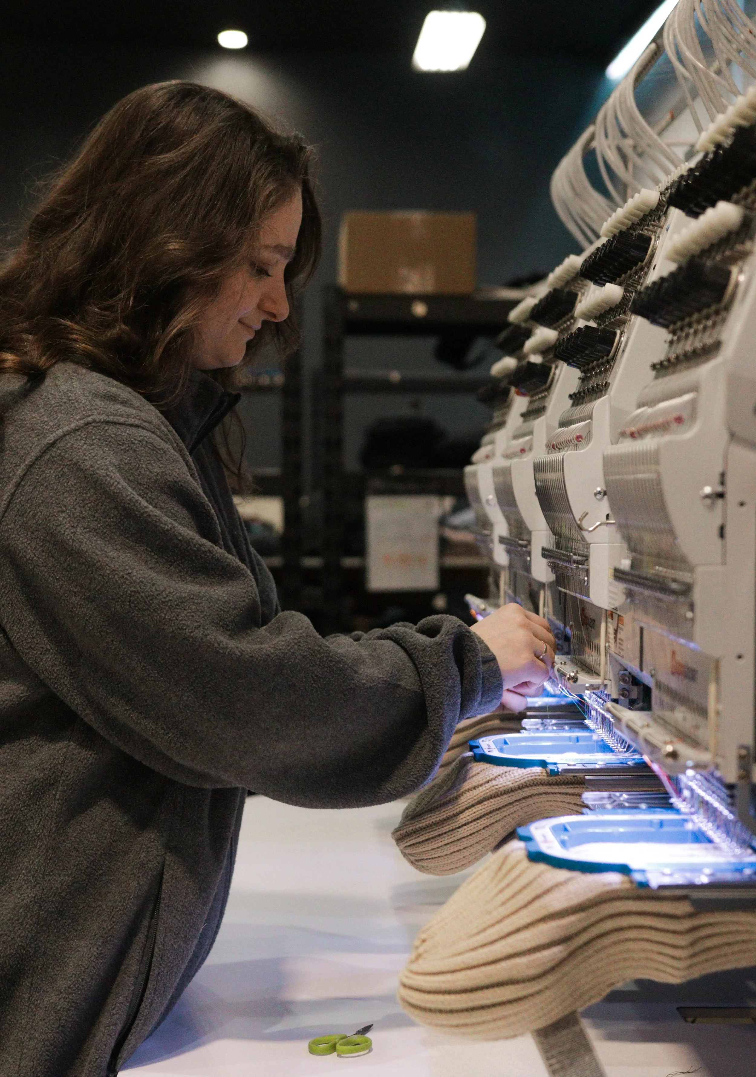 A woman working on embroidery machines in a factory or workshop setting, wearing a grey fleece jacket and focused on her work.