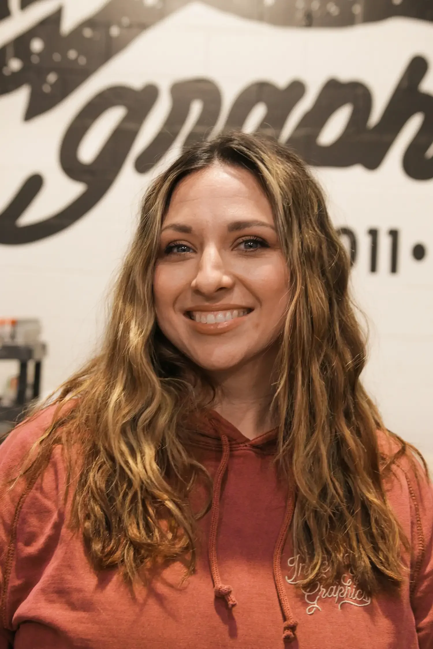 A woman with long wavy brown hair smiling in front of a wall with black text that is partially visible.