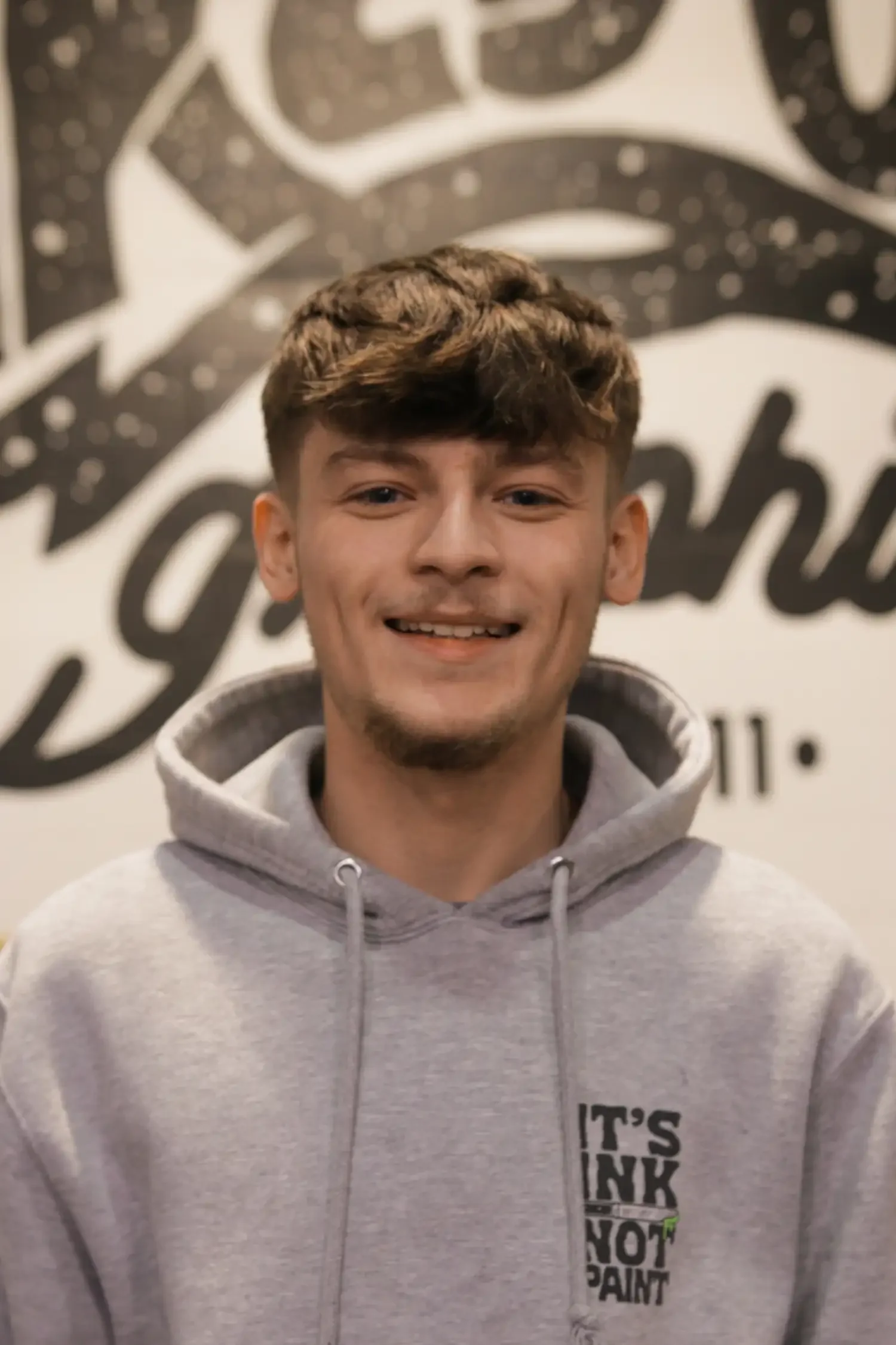 A young man with light brown hair smiling, wearing a gray hoodie with text in black that reads 'IT'S INK NOT PAINT,' standing in front of a black and white mural or wall art background.
