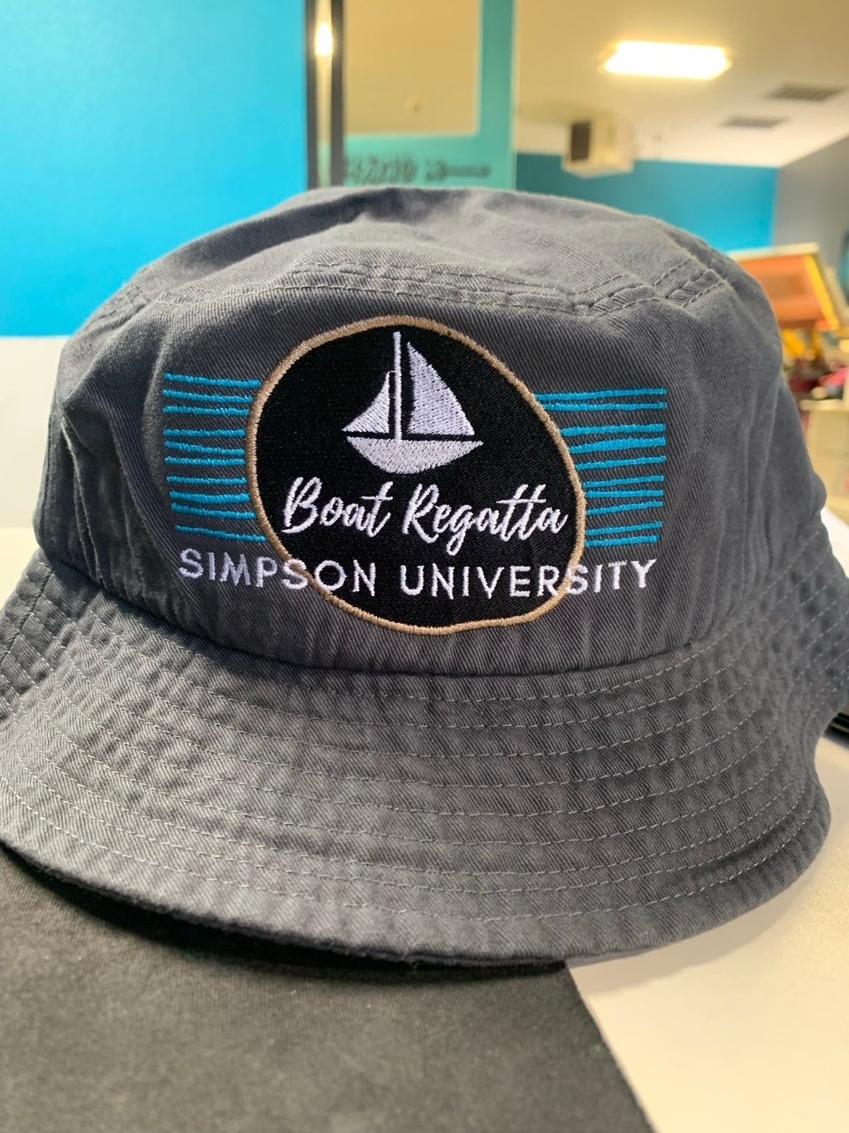 Gray bucket hat with embroidered logo of a sailboat, text reading 'Boat Regatta,' and 'SIMPSON UNIVERSITY'.