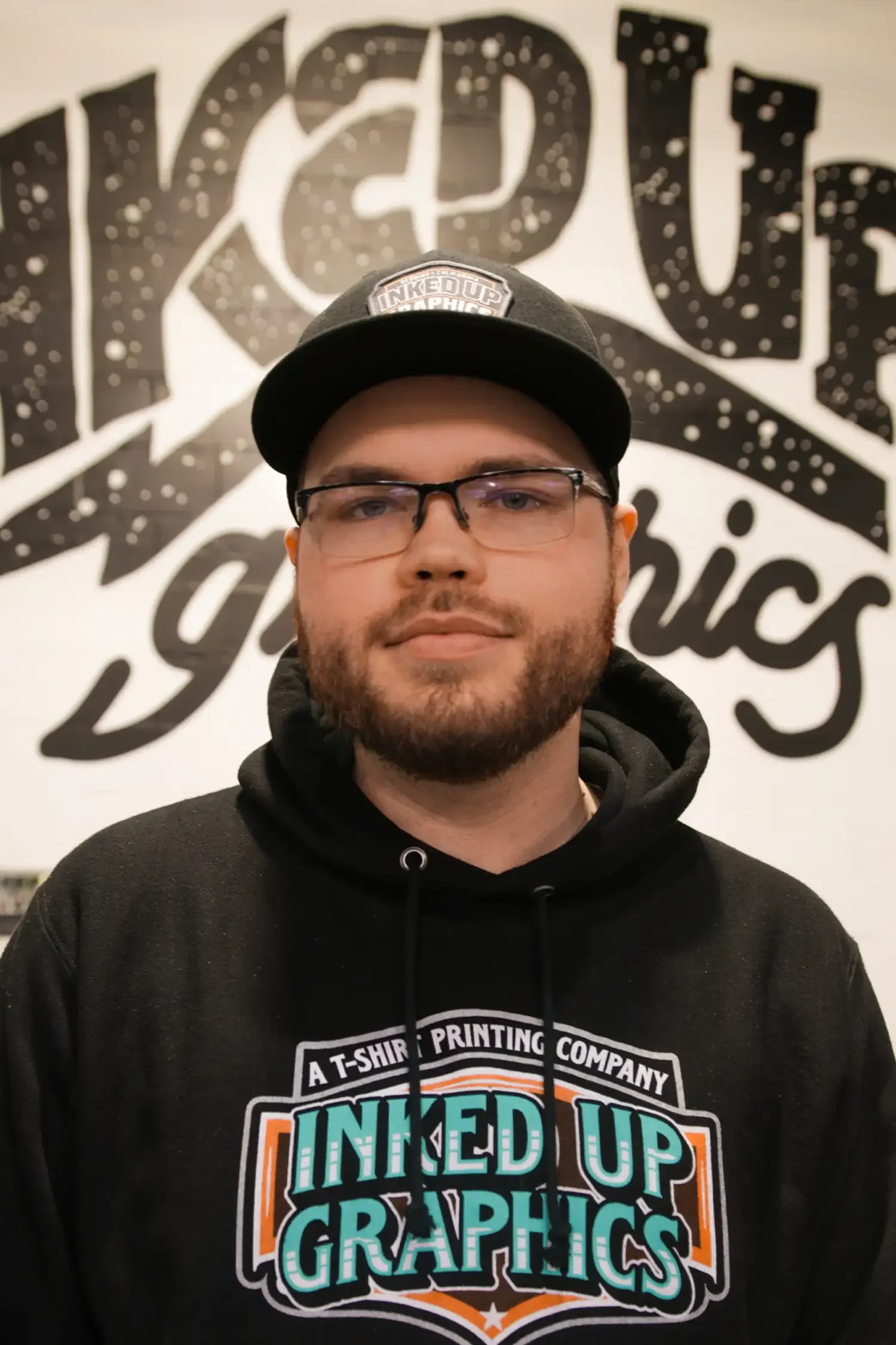 A young man with glasses and a beard wearing a black hoodie and a black cap with the logo 'Inked Up Graphics' stands in front of a wall with a large black and white sign that says 'Inked Up Graphics'.