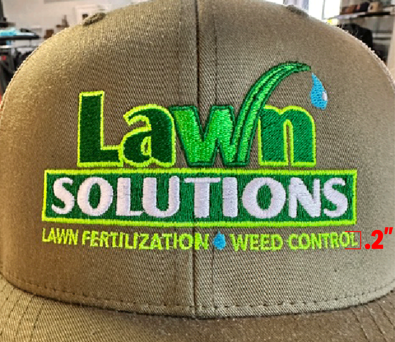 Close-up of a beige trucker hat with an embroidered logo for 'Lawn Solutions' in green, white, and yellow text, with additional text 'Lawn Fertilization' and 'Weed Control' in yellow and green, and a small blue water droplet graphic.