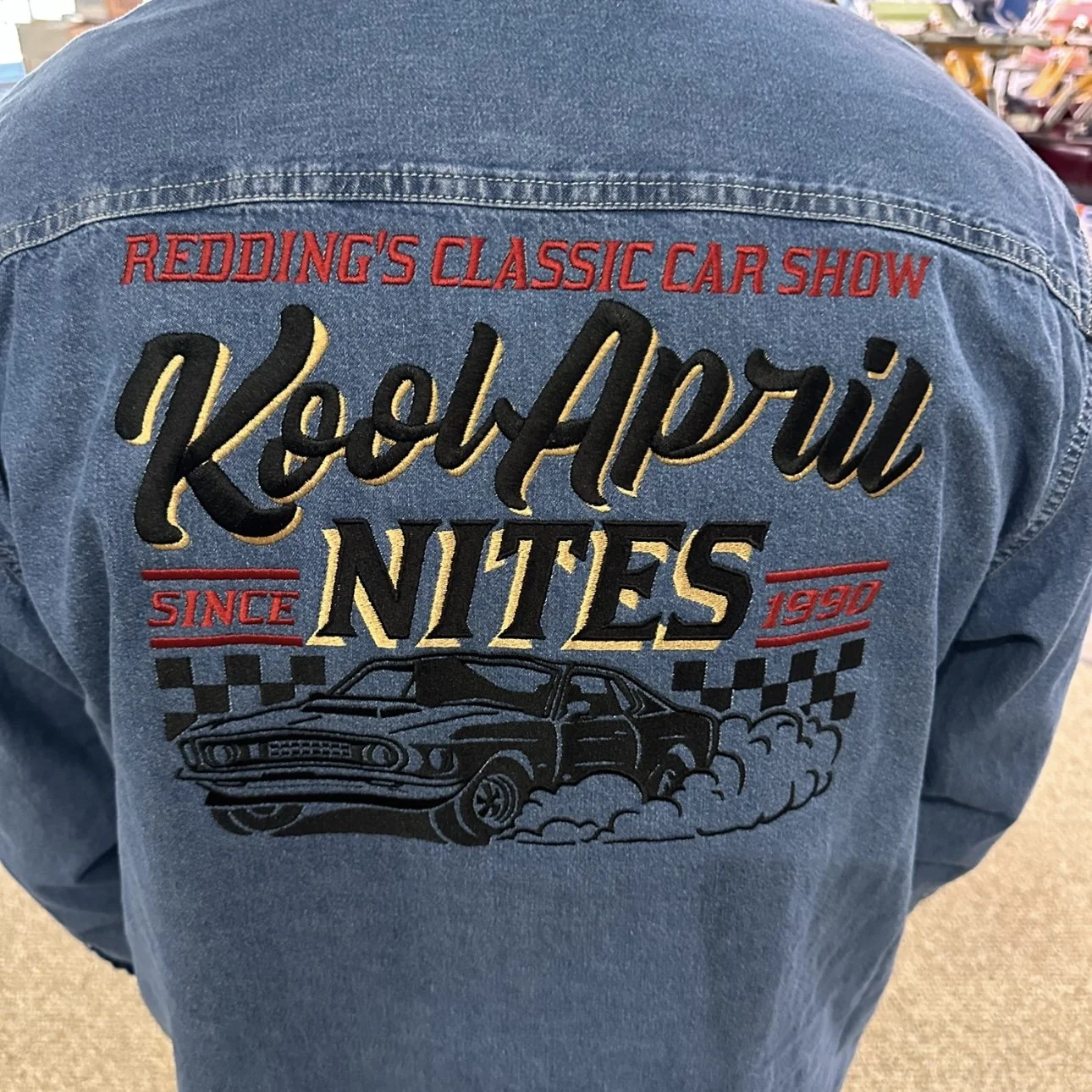 Back of a denim jacket with embroidered text and a vintage car graphic. The embroidered text reads 'Redding's Classic Car Show', 'KoolAori Nites', 'Since 1950', with a black car illustration below, surrounded by checkered flags.