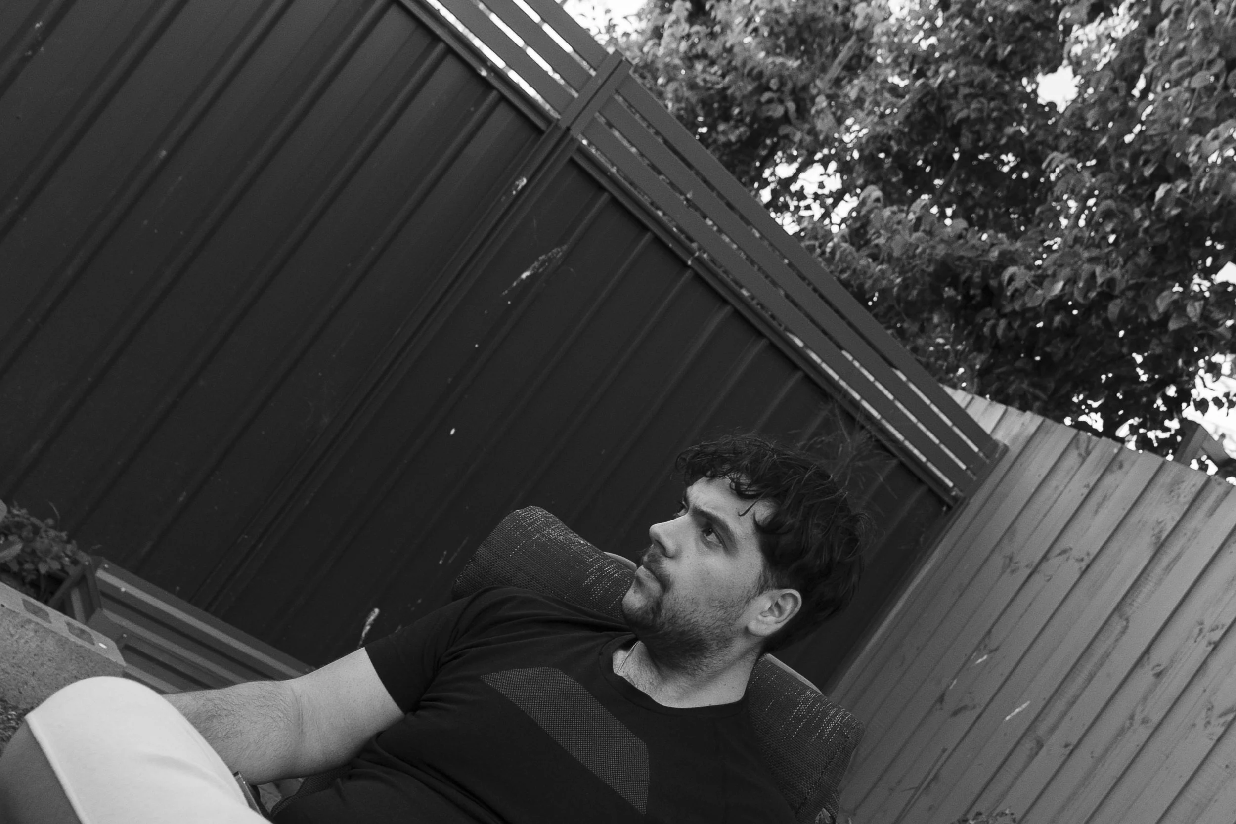 A young man with dark, curly hair and a beard, sitting outdoors in a chair, looking to his right with a thoughtful expression. He is near a wooden fence and some greenery.