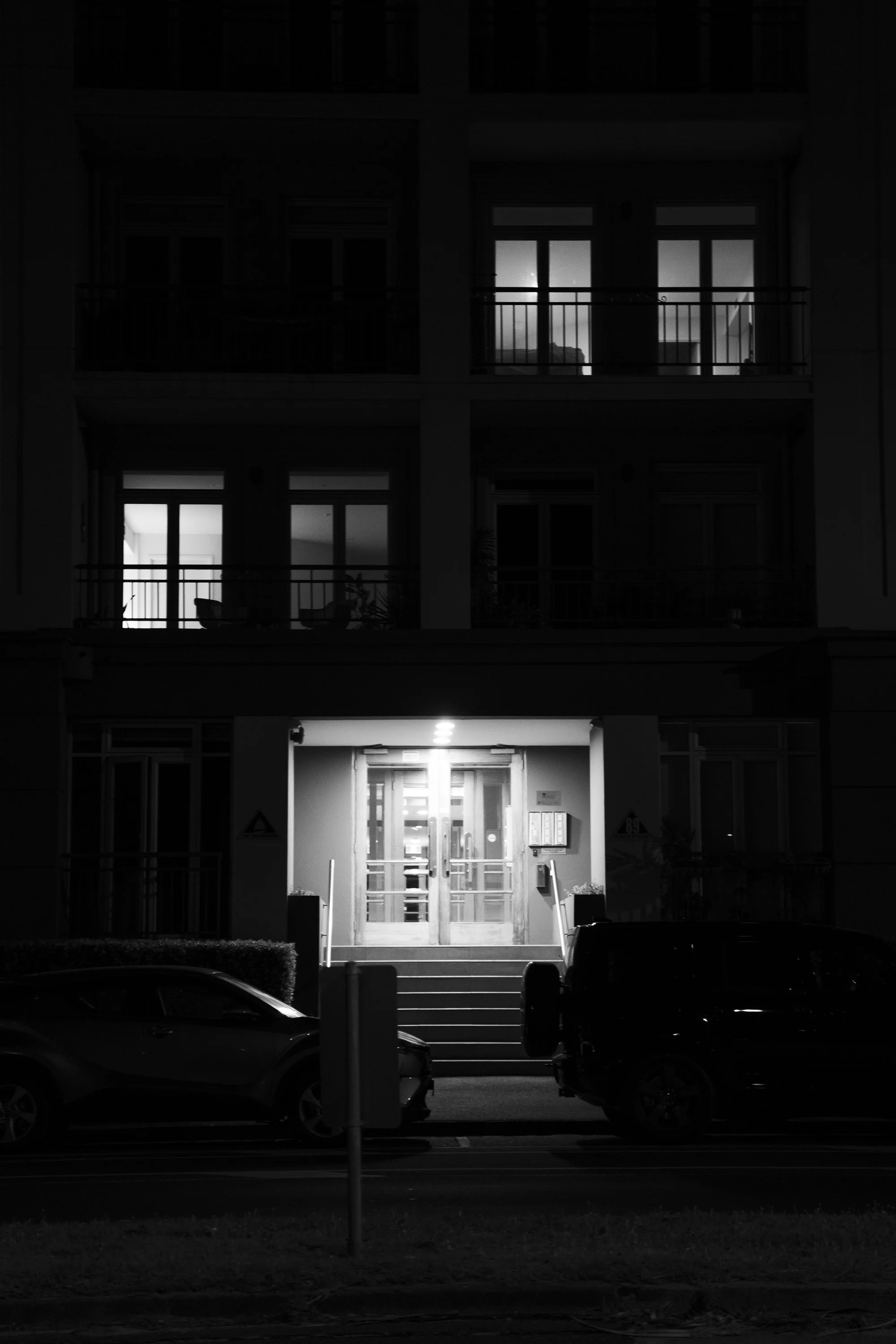 Nighttime view of an apartment building with illuminated entrance and two parked cars in front.