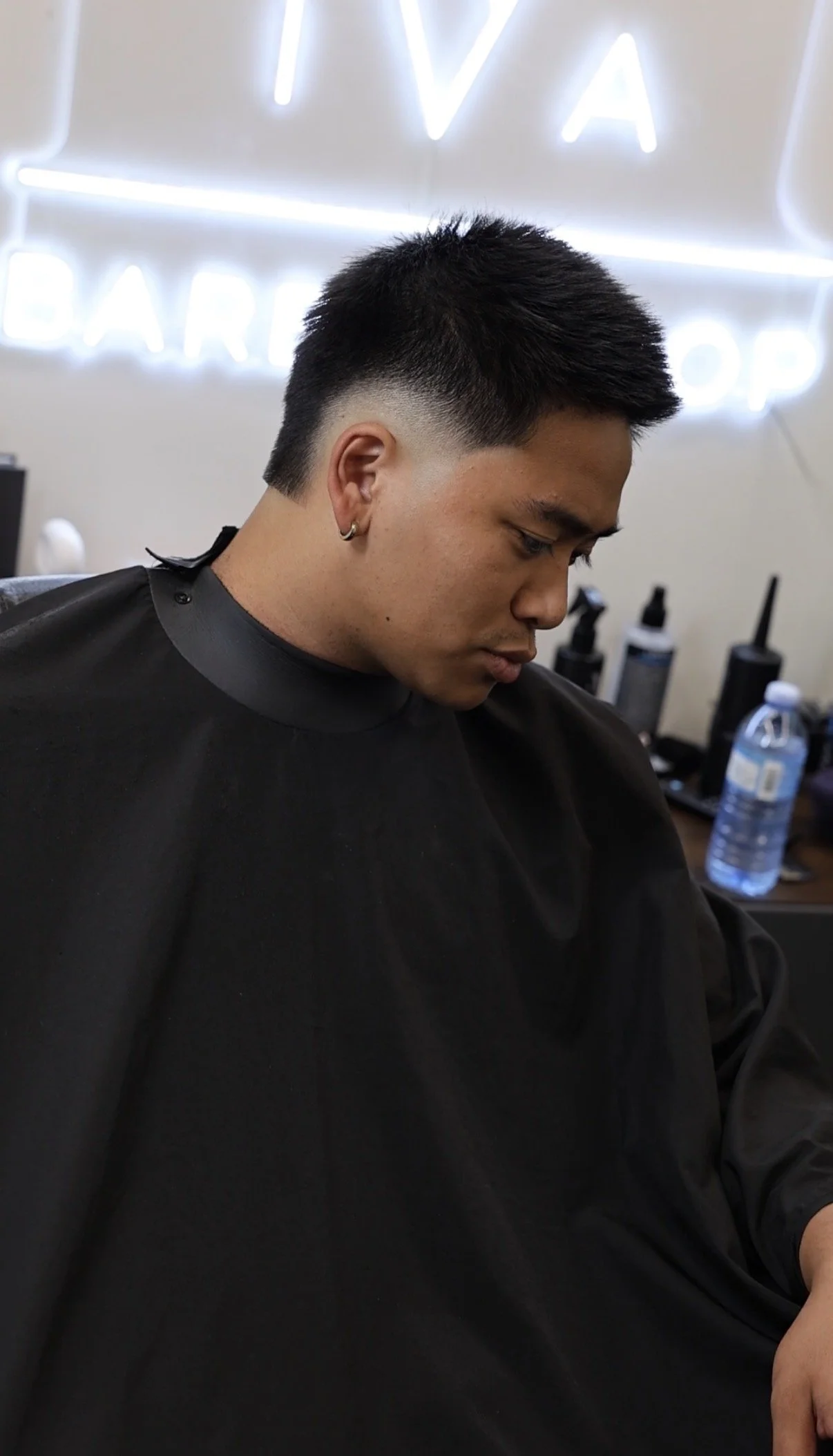 Young man with short, textured haircut sitting in barber chair, wearing black cape, looking down, with barber supplies and a water bottle in the background, and neon sign reading 'VIVA BARBER SHOP' on the wall.