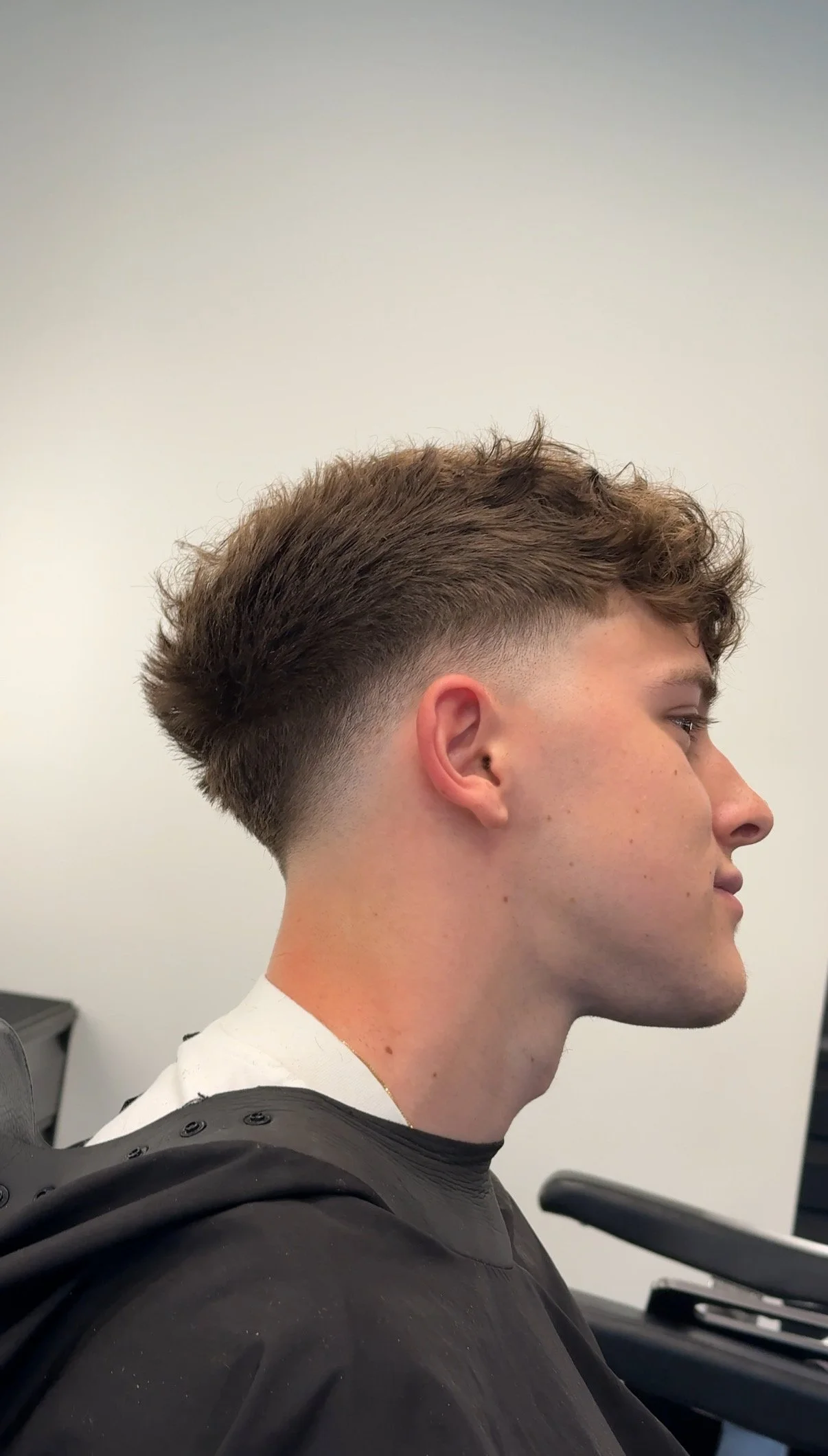 Side profile of a young man with a modern fade haircut, wearing a black salon cape, sitting in a barbershop chair.