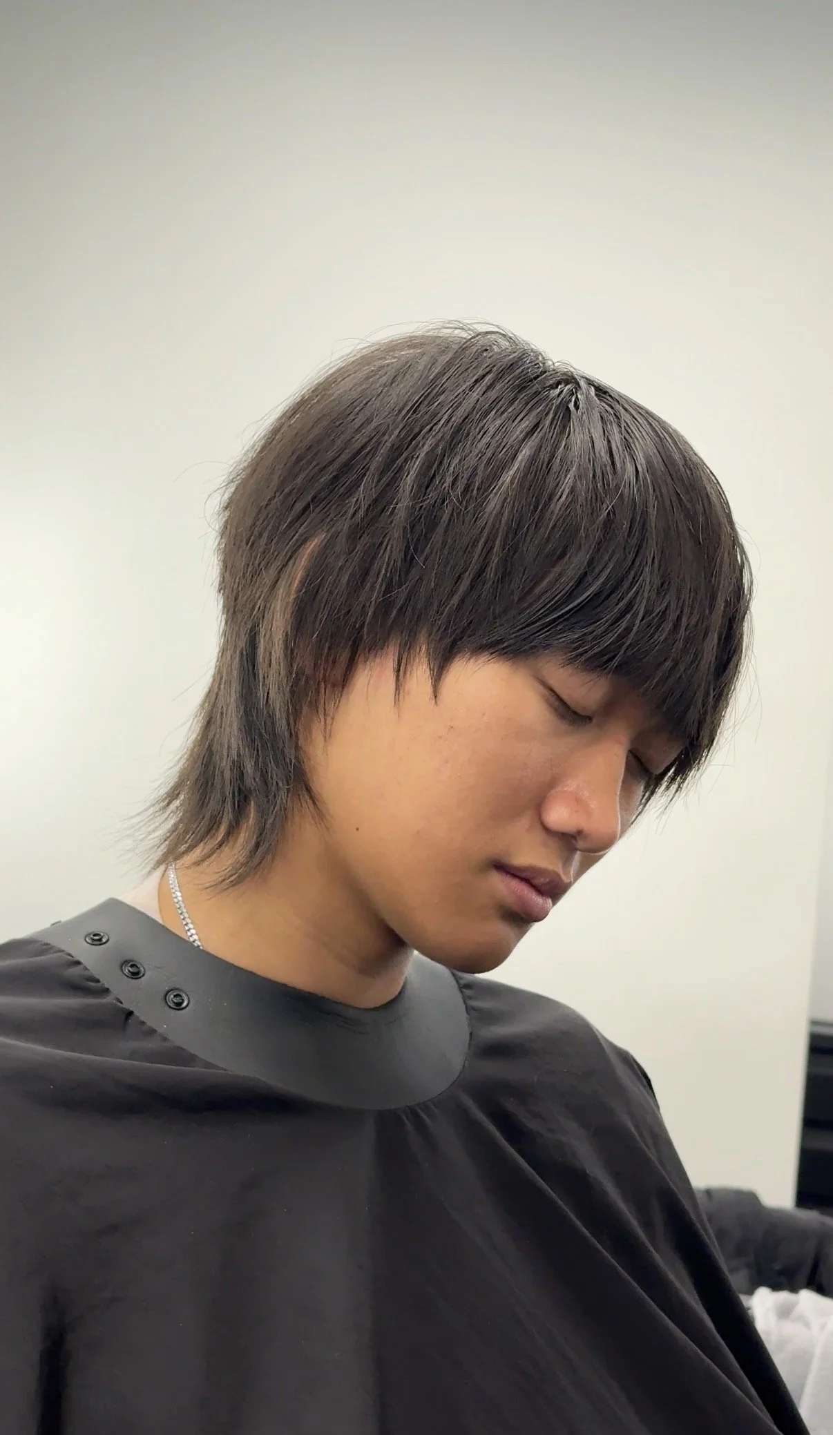 A young man with straight, layered dark hair, closed eyes, wearing a black haircut cape in a salon.