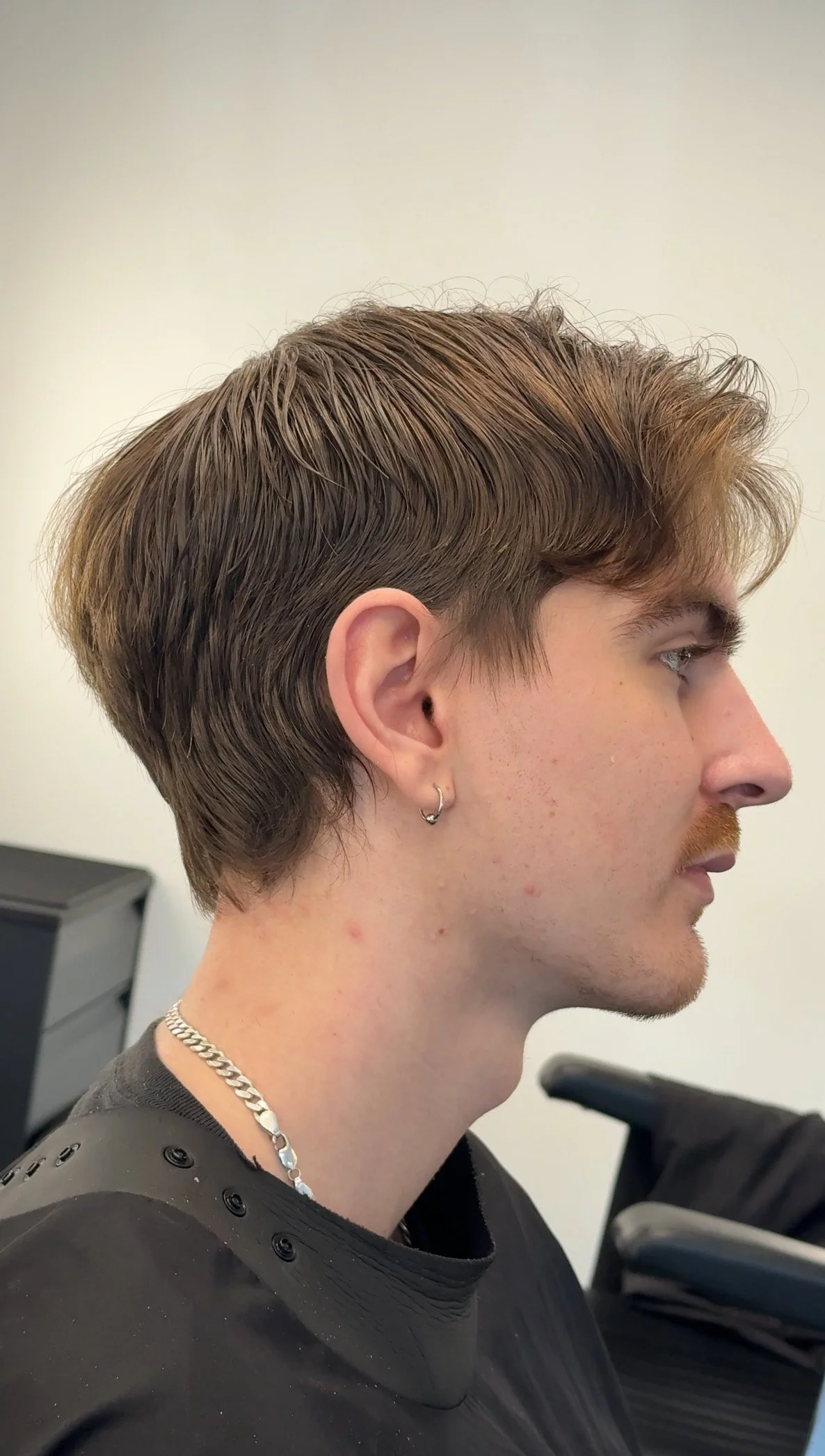 Side profile of a young man with light brown hair, ear piercing, wearing a black shirt, a chain necklace, and sitting in a chair.