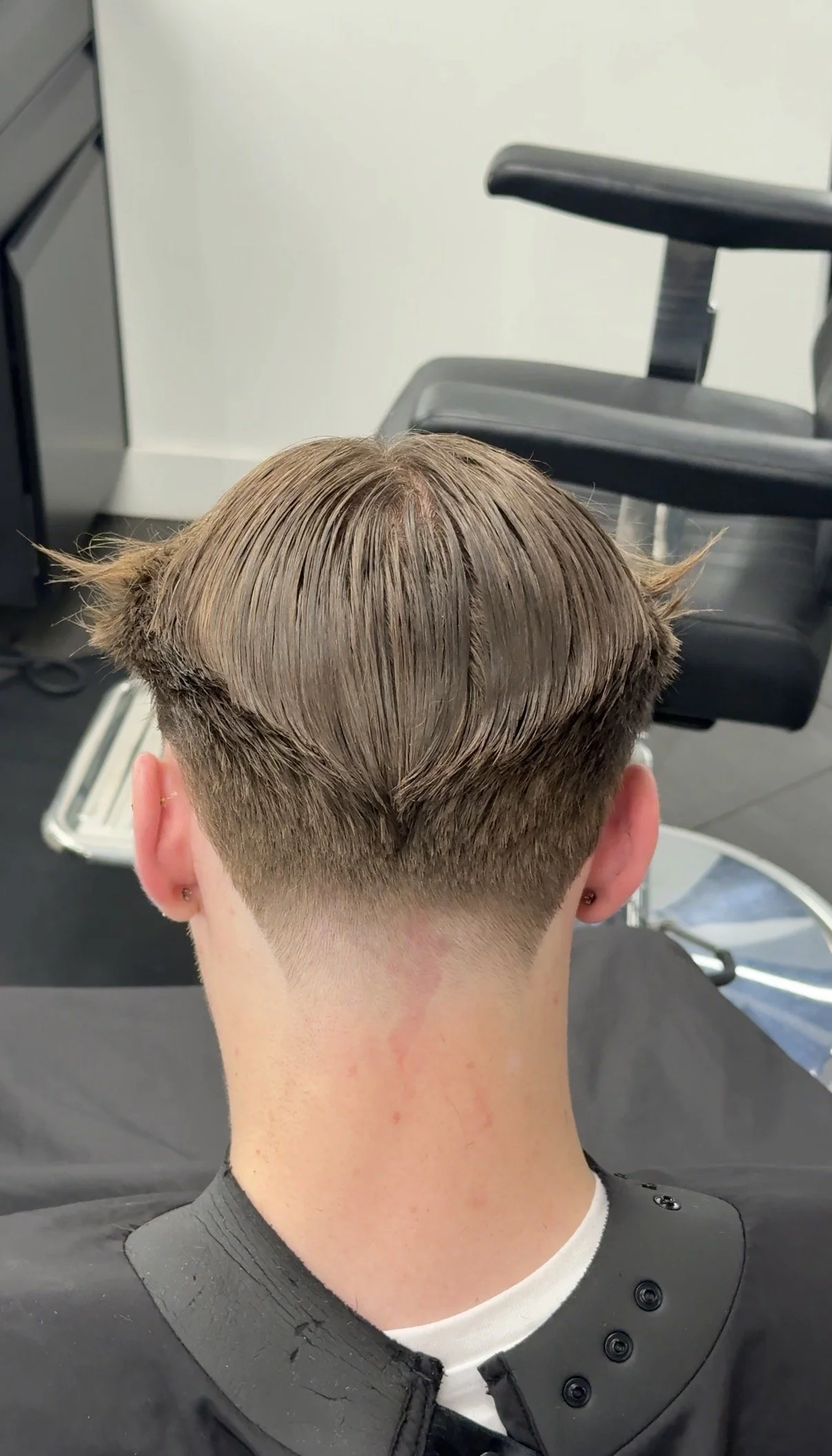 Back view of a young person's freshly cut hairstyle, with short sides and longer top hair, in a barber shop.