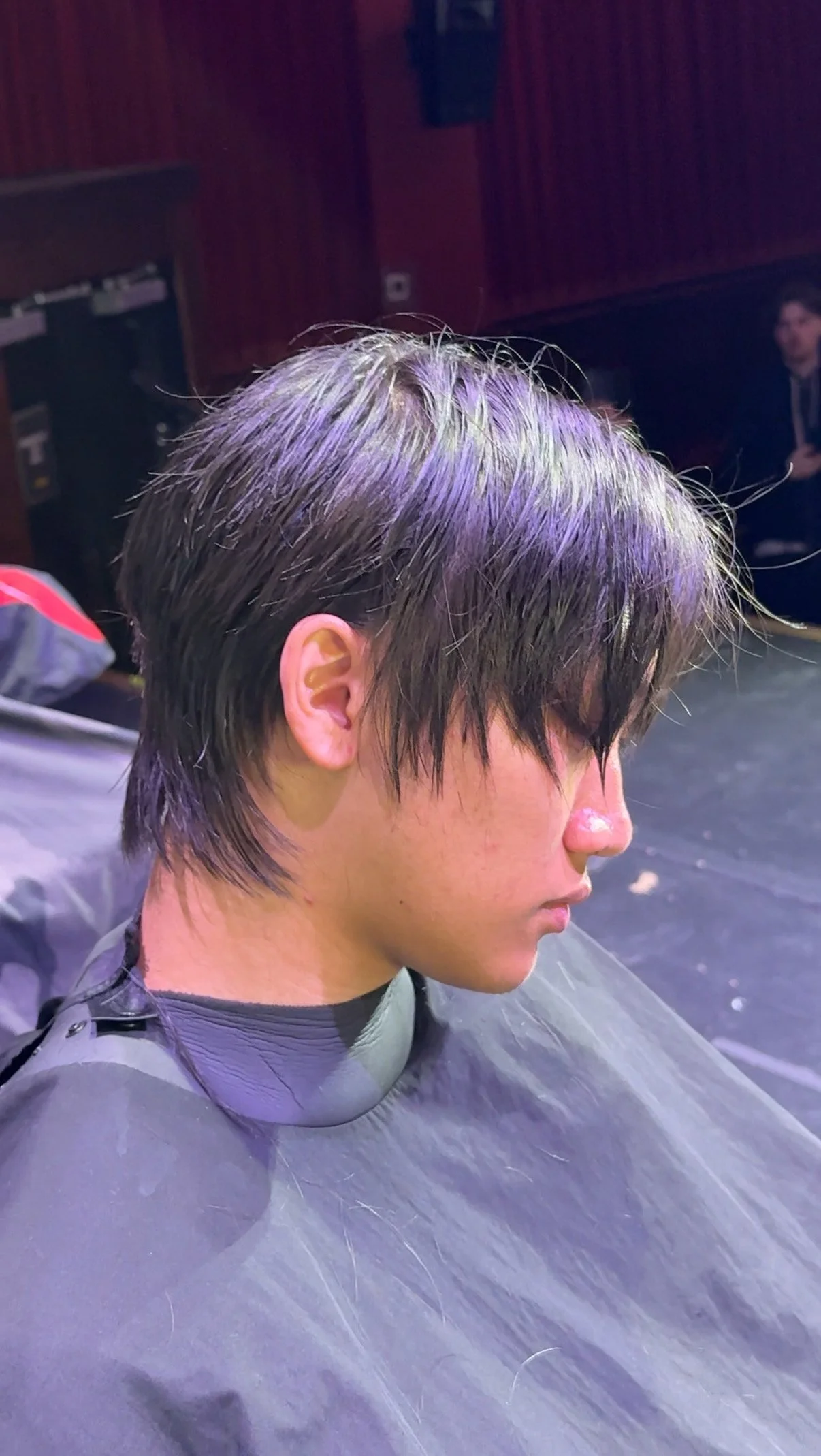 Side view of a young person with wet, short, dark hair sitting in a chair, possibly in a salon or barbershop, with a dark background and another person in the distance.