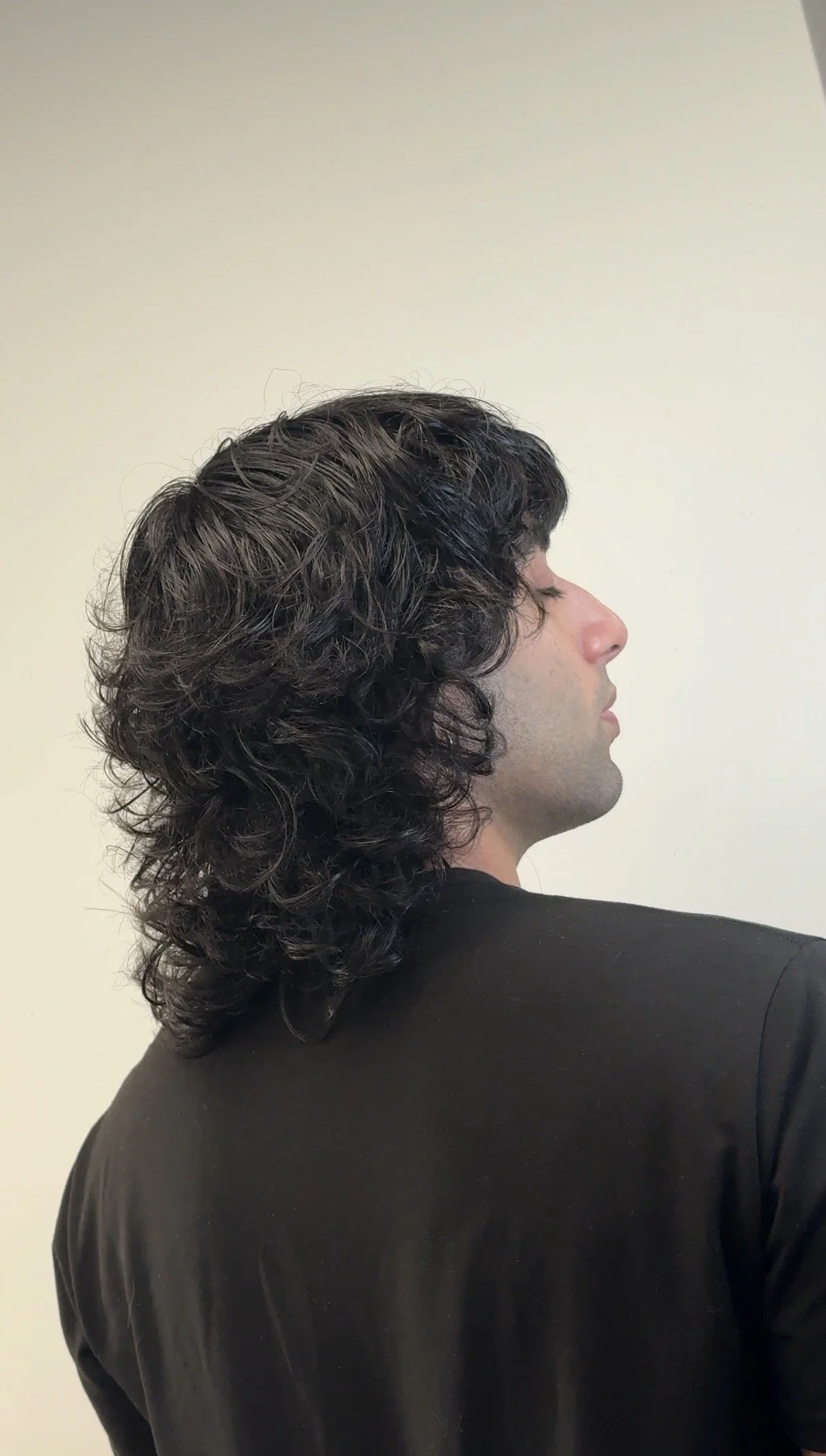 Profile of a man with long, curly black hair wearing a black shirt against a plain light-colored wall.