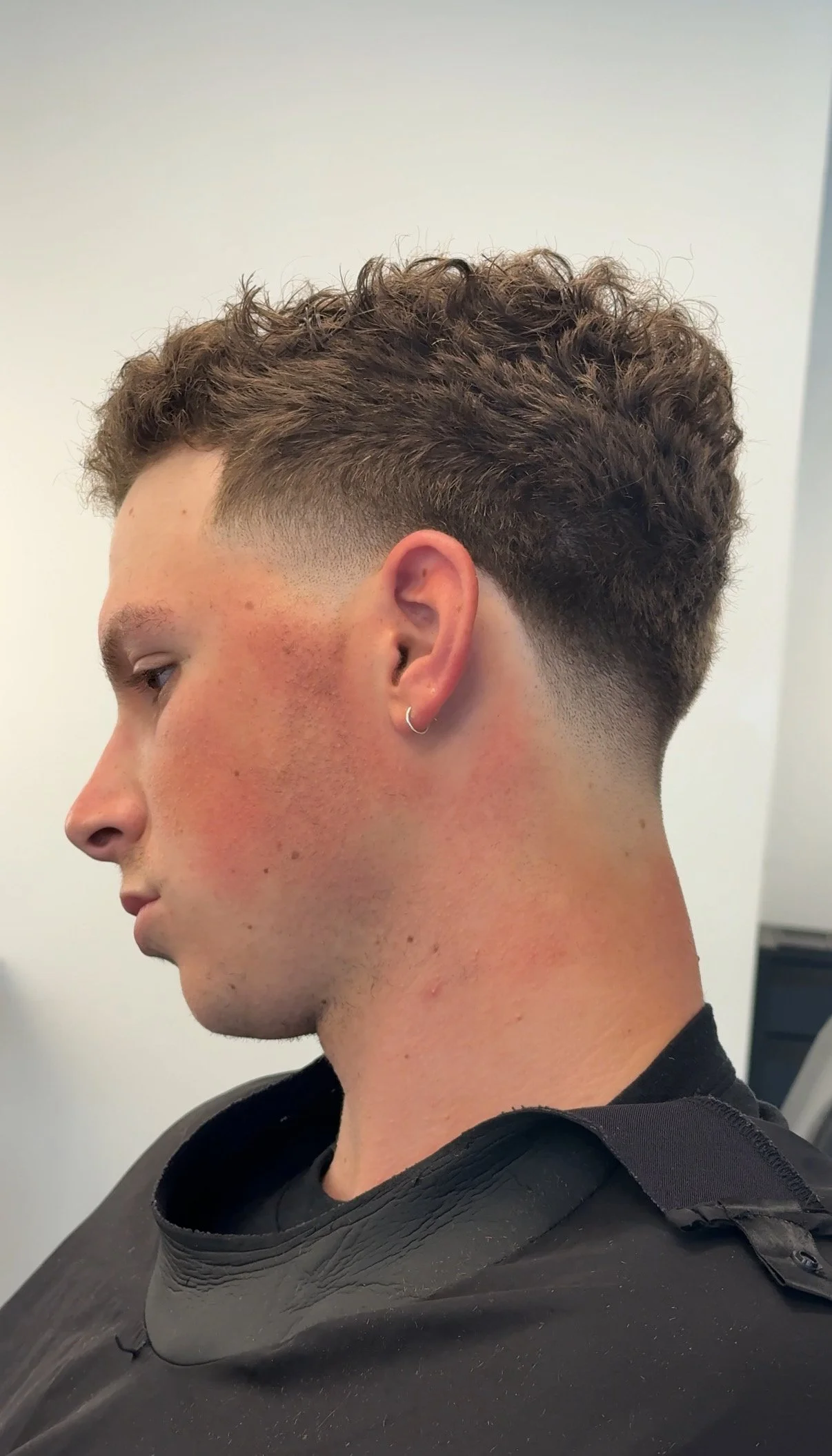 Side view of a young man with a fresh haircut, featuring curly hair on top and a sharp fade on the sides, wearing a black shirt and a silver hoop earring.