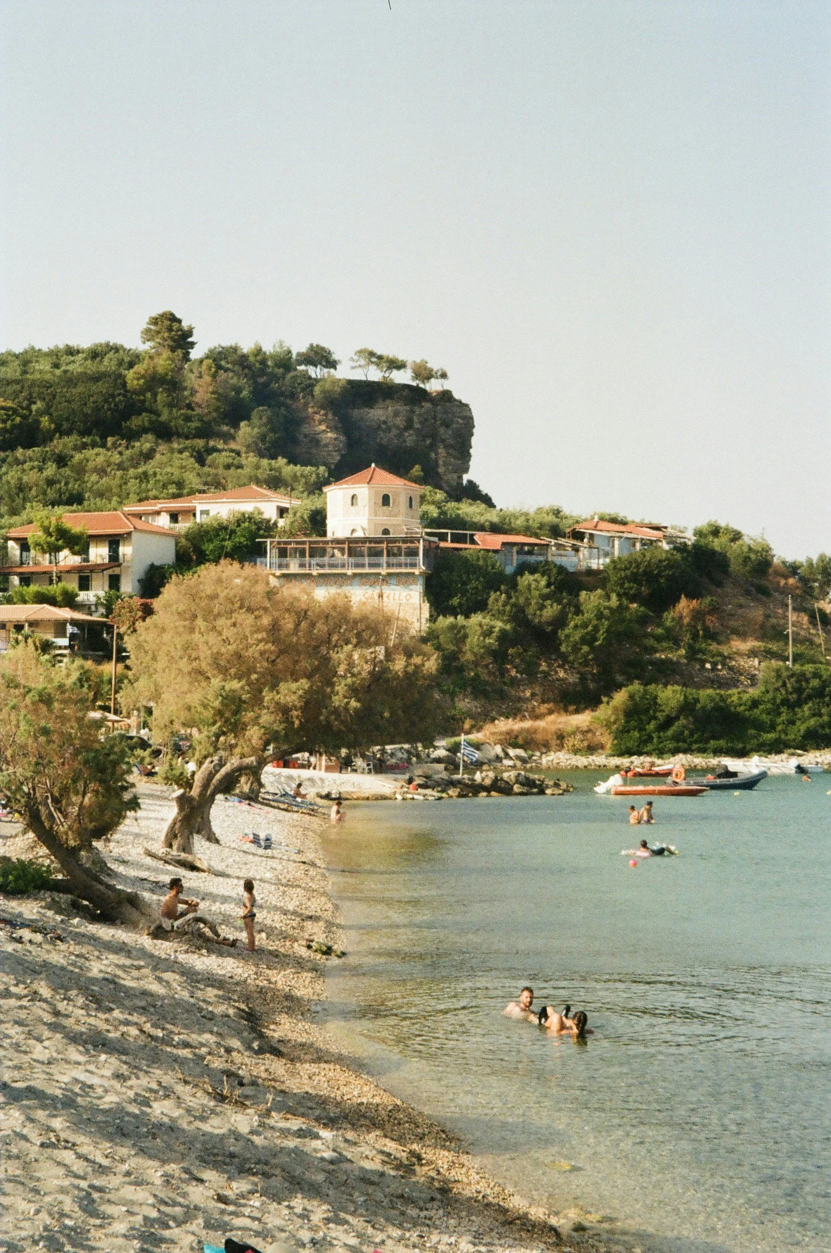 A beach scene with people swimming and relaxing in the water and on the sandy shore. There are trees along the shore and buildings on a hill in the background. Several boats are floating in the water near the beach.
