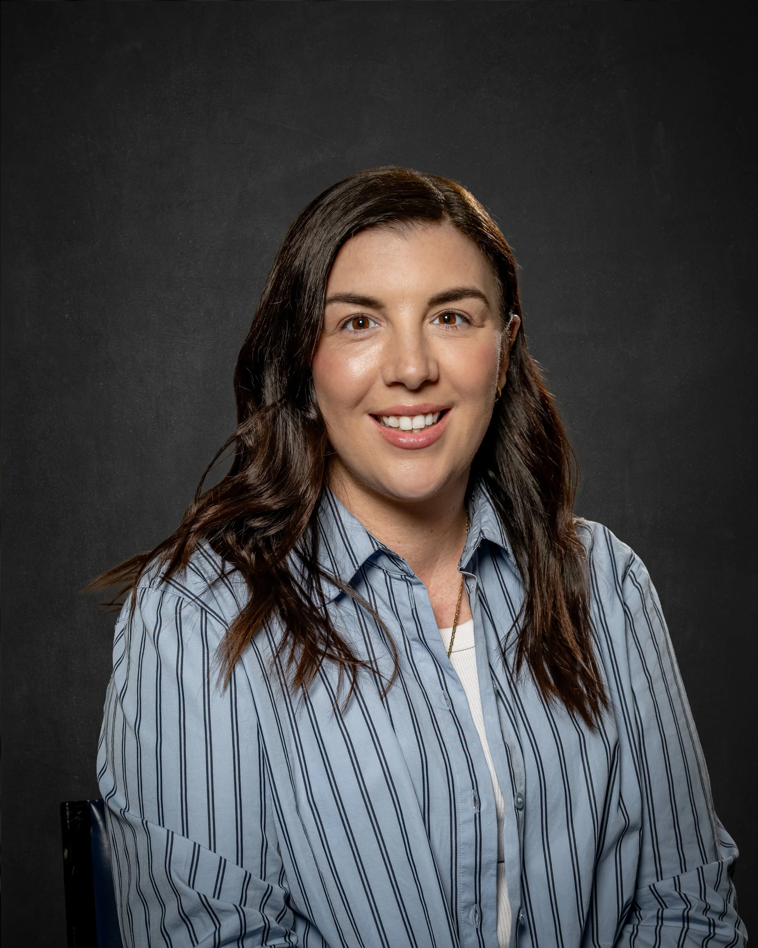 A woman with long brown hair, wearing a blue and white striped button-up shirt, sitting on a black chair against a dark background, smiling with visible teeth.