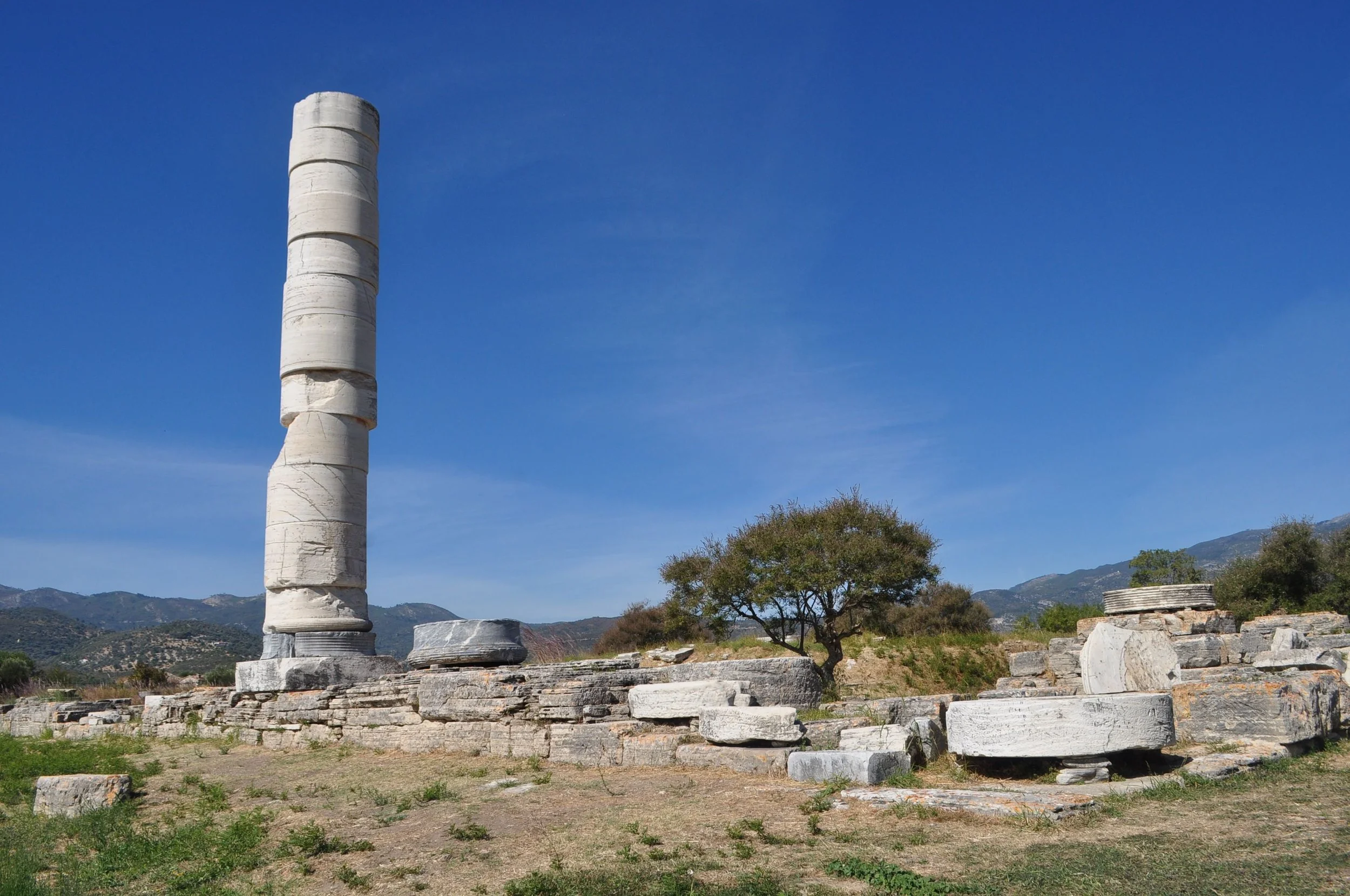 Ancient Greek ruins with a large, white marble column standing upright among scattered stone blocks and ruins. There is a small round structure in the background, trees, and mountains under a clear blue sky.