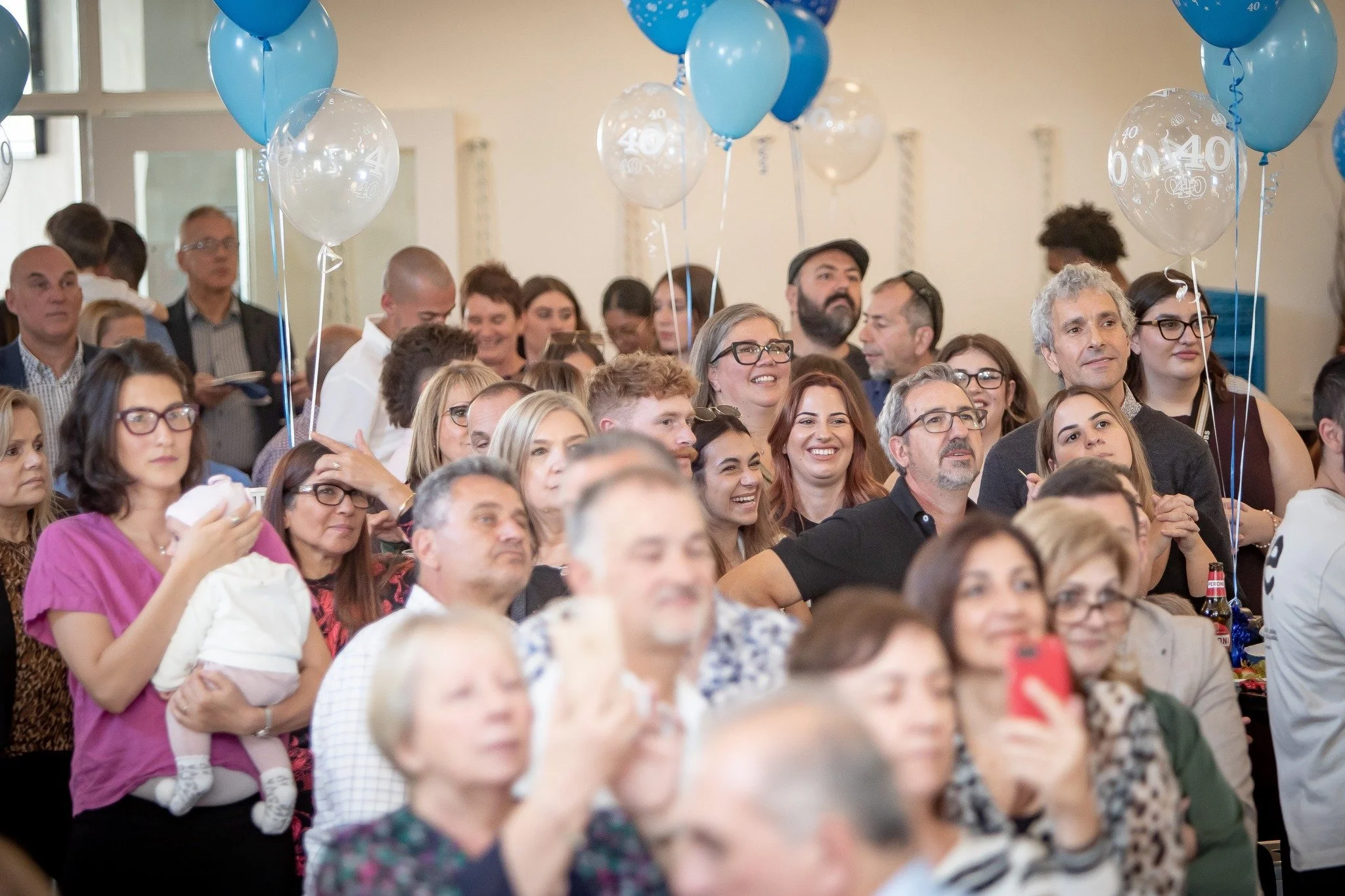 A large group of diverse people gathered in a room, celebrating a 40th anniversary with blue and transparent balloons that have '40' printed on them.