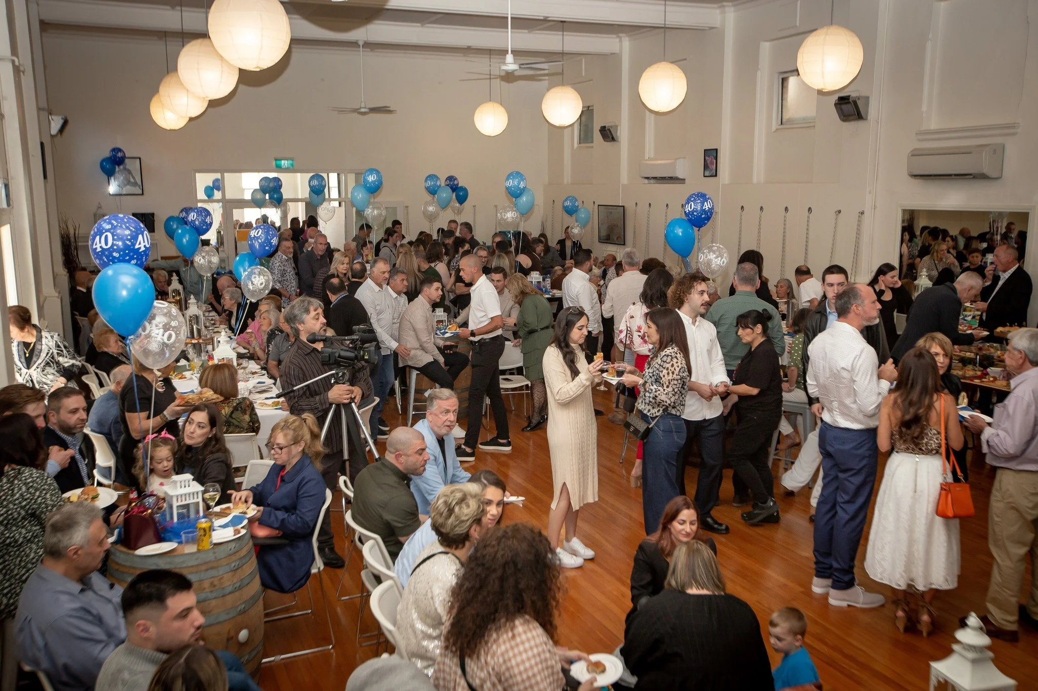 A large indoor celebration with many people gathered around tables, some standing, some seated, celebrating a 40th anniversary or birthday with blue and silver balloons that have the number 40 on them. The room has hanging paper lanterns, a wooden floor, and white walls.