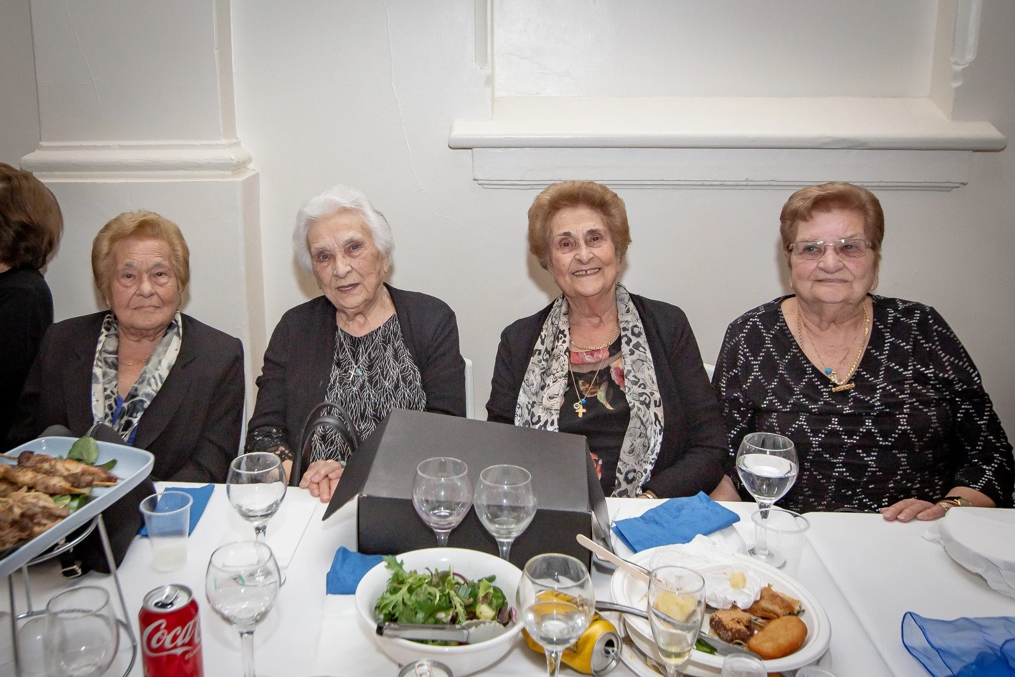 Four elderly women sitting at a table during a social gathering, with plates of food and drinks, in a white-walled room.