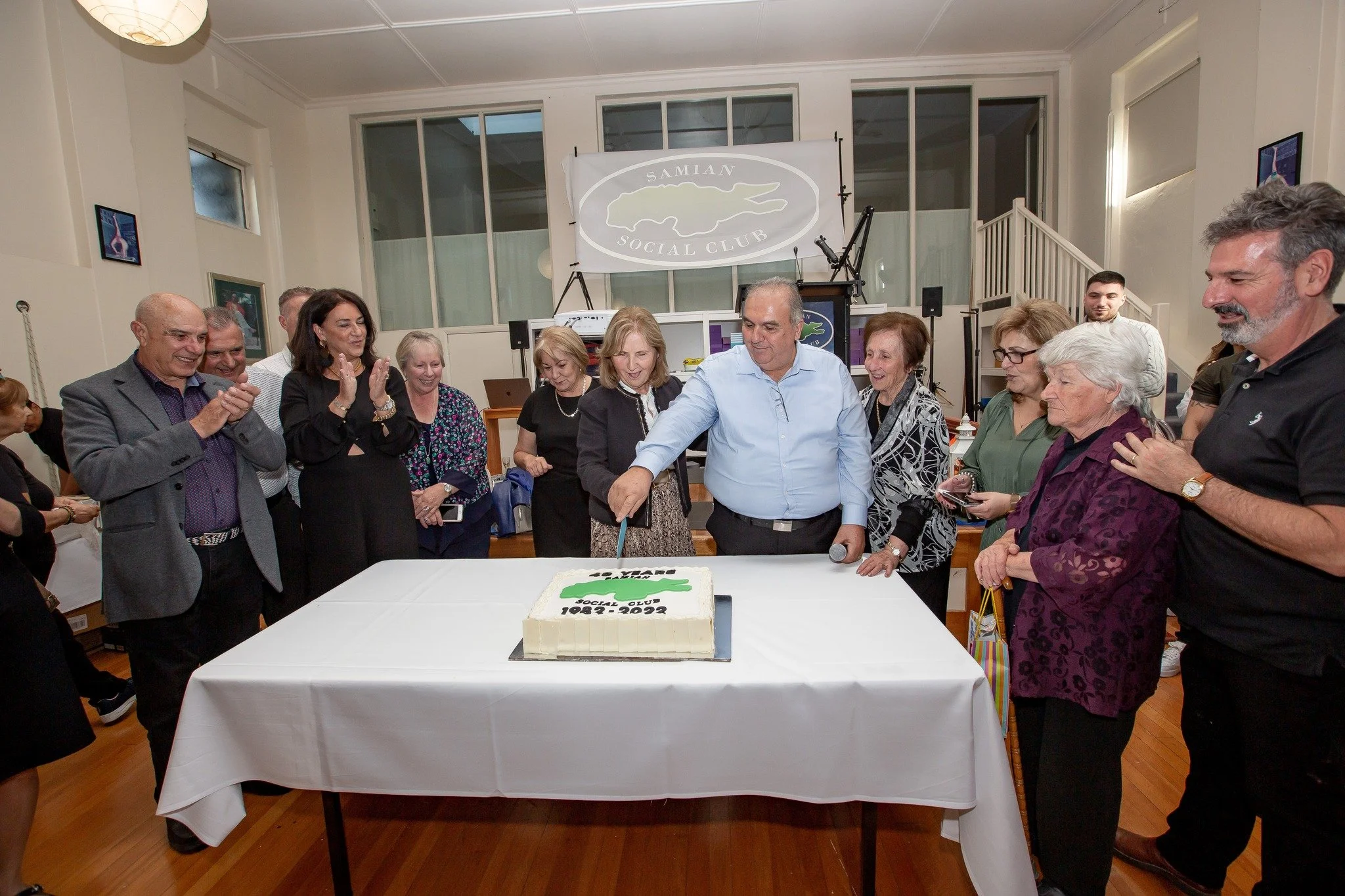 A group of people gathered around a table with a cake that reads 'SAMIAN SOCIAL CLUB 1972-2022.' They are celebrating and smiling, with some clapping.
