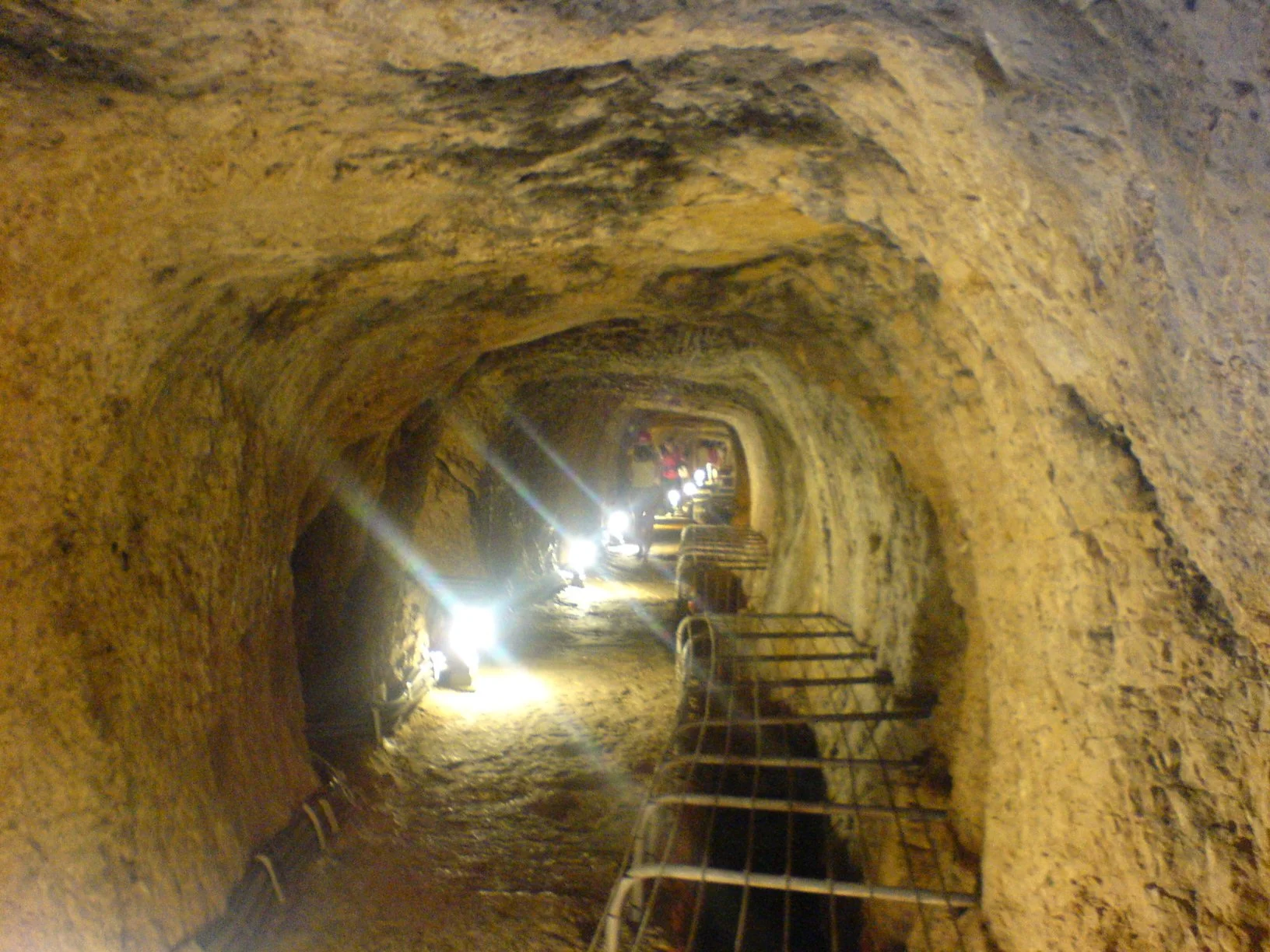 Underground tunnel with rough, uneven stone walls and ceiling, illuminated by bright lights along the path, with metal railings and a walkway.