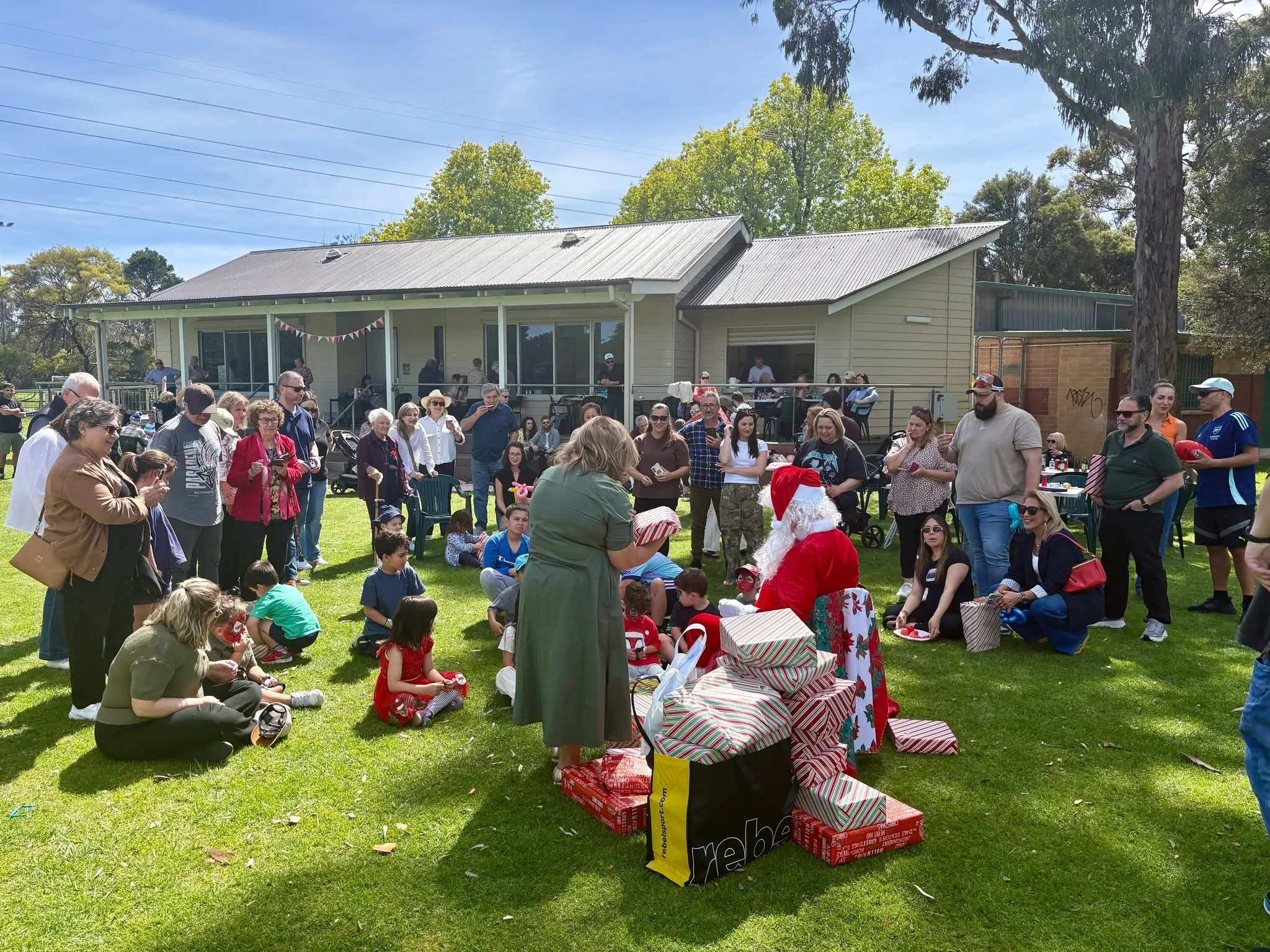 A large gathering of people at an outdoor event, involving children and adults, with some sitting on the grass, and others standing, facing towards a person dressed as Santa Claus, who is seated among wrapped presents. There are trees, a house-style building, and string lights in the background under a sunny sky.